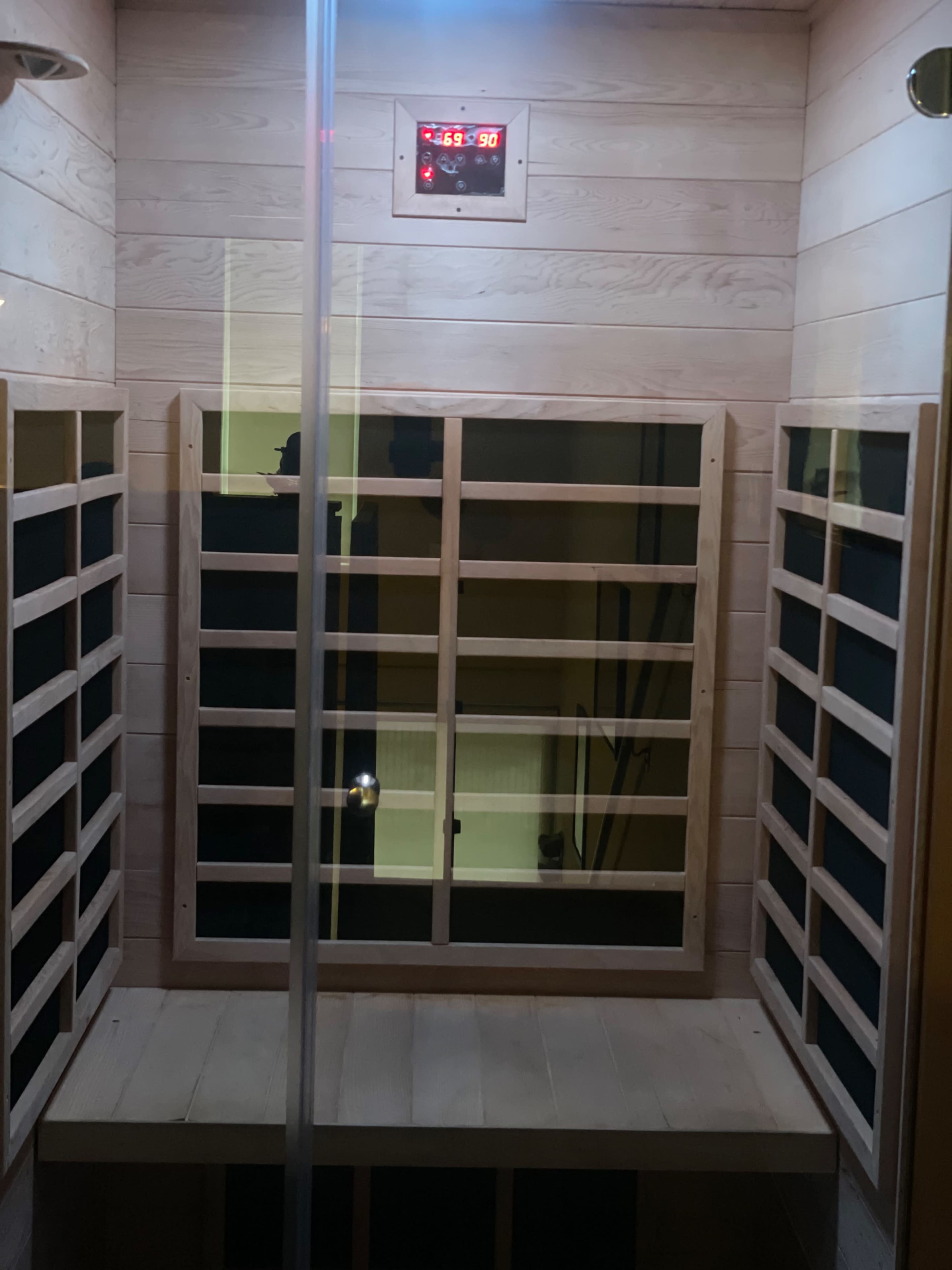 The image shows an infrared sauna with wooden paneling, benches, and a digital timer mounted on the wall.