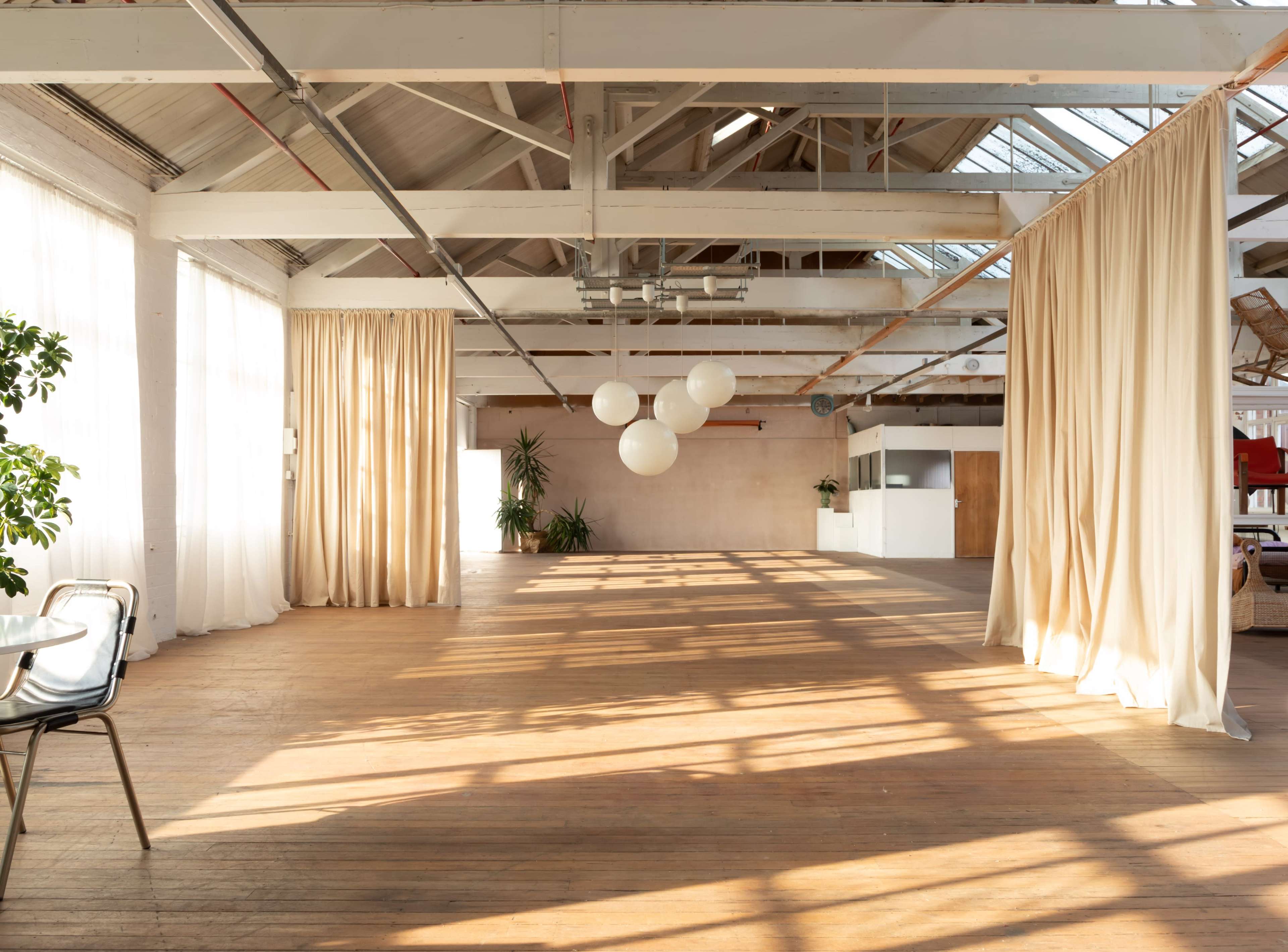 The image shows a spacious interior with wooden flooring, natural light streaming in from large windows, and light-colored curtains separating sections of the room.