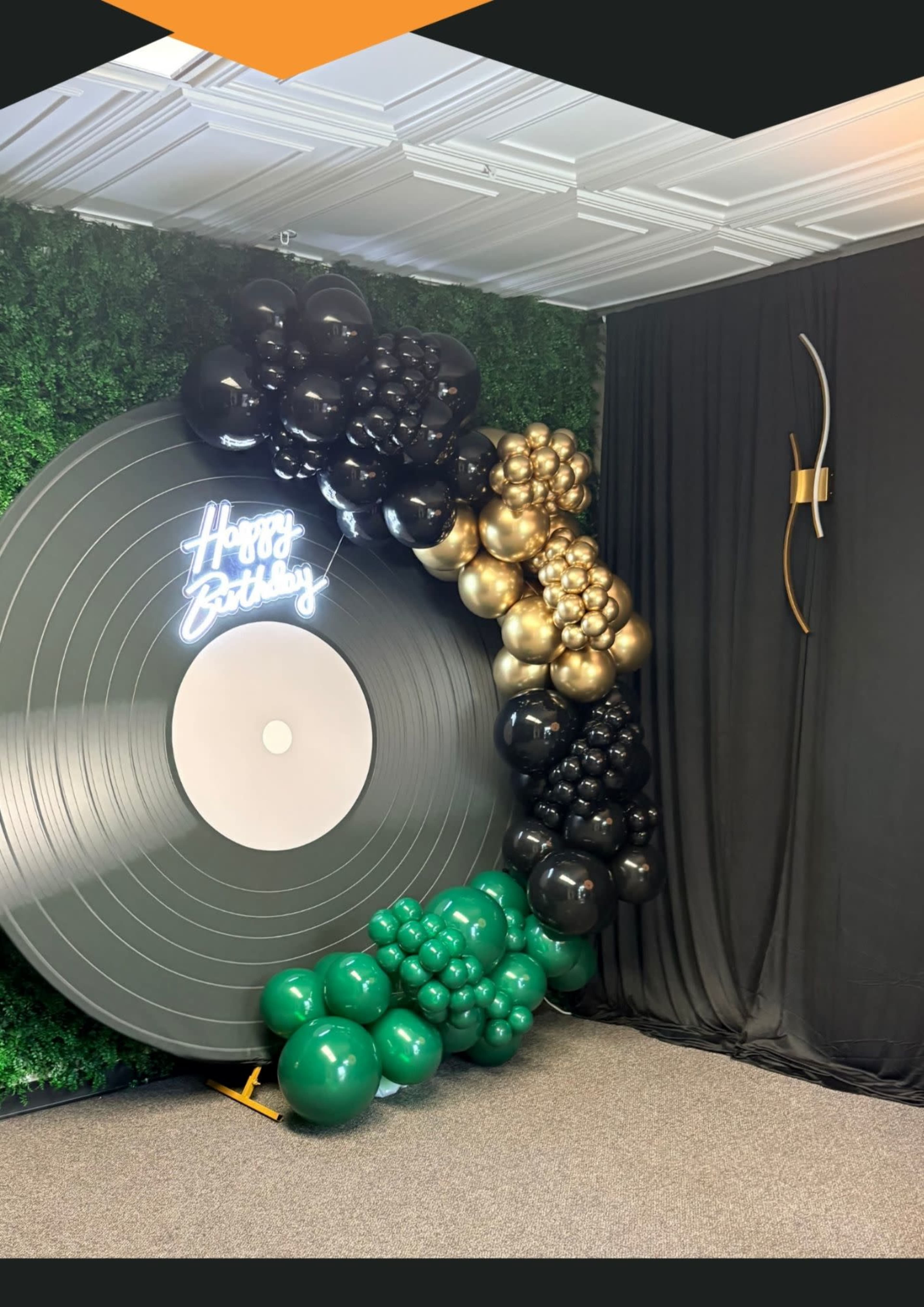 The image shows a birthday decoration featuring a large vinyl record backdrop surrounded by an arrangement of balloons in black, gold, and green.