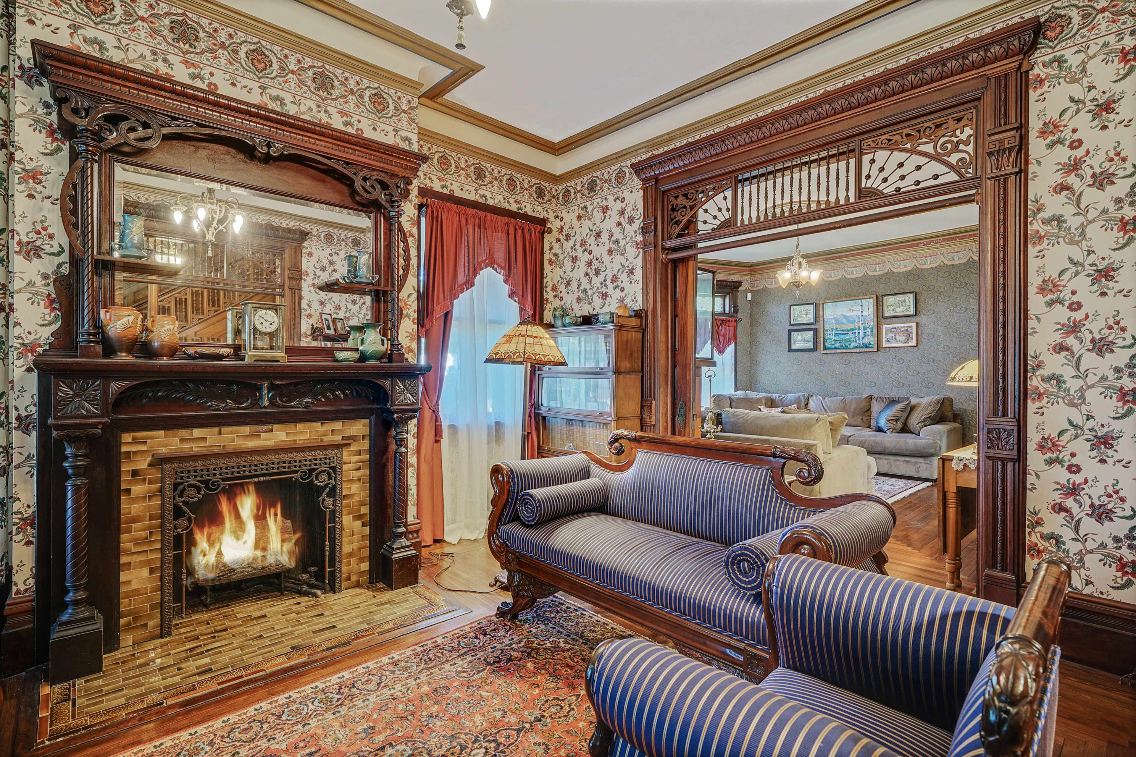 A vintage living room with a patterned wallpaper, an ornate wooden fireplace, and a blue-striped sofa facing a mirror.