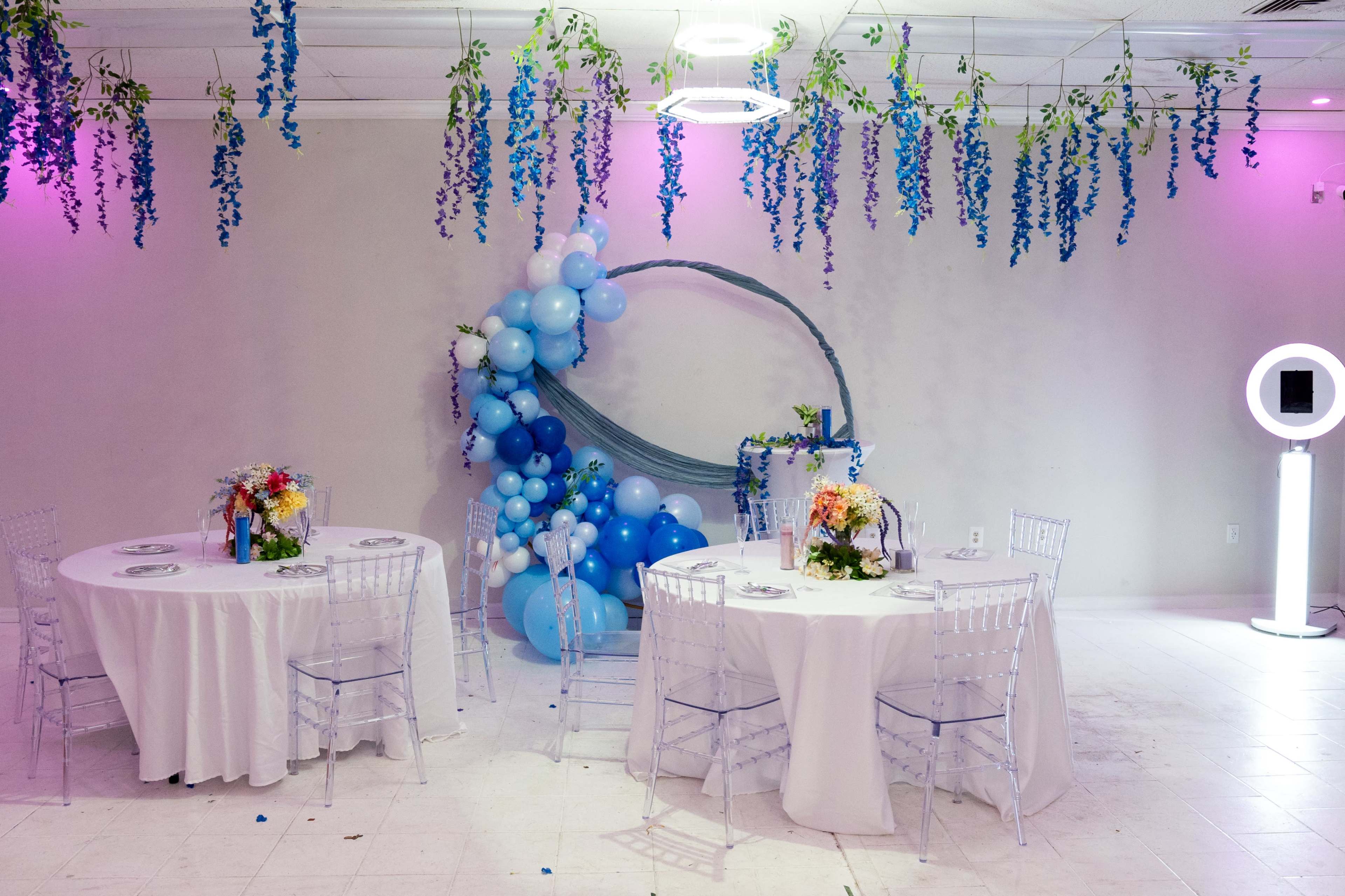 The image shows a decorated event space with two round tables covered in white tablecloths, surrounded by blue and white balloon arrangements and suspended floral decorations.