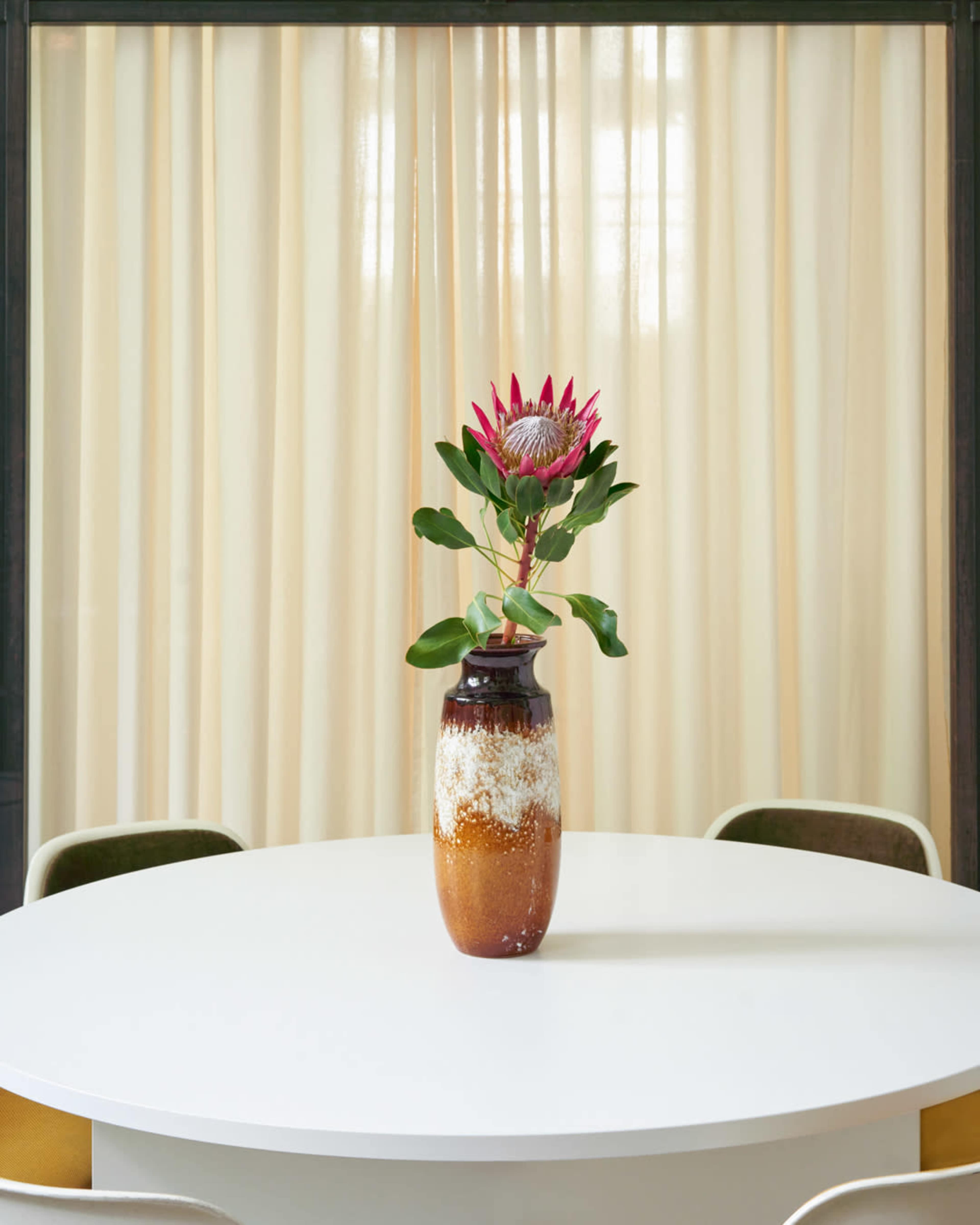 A large vase with a pink flower and green leaves sits on a round white table in front of sheer curtains.