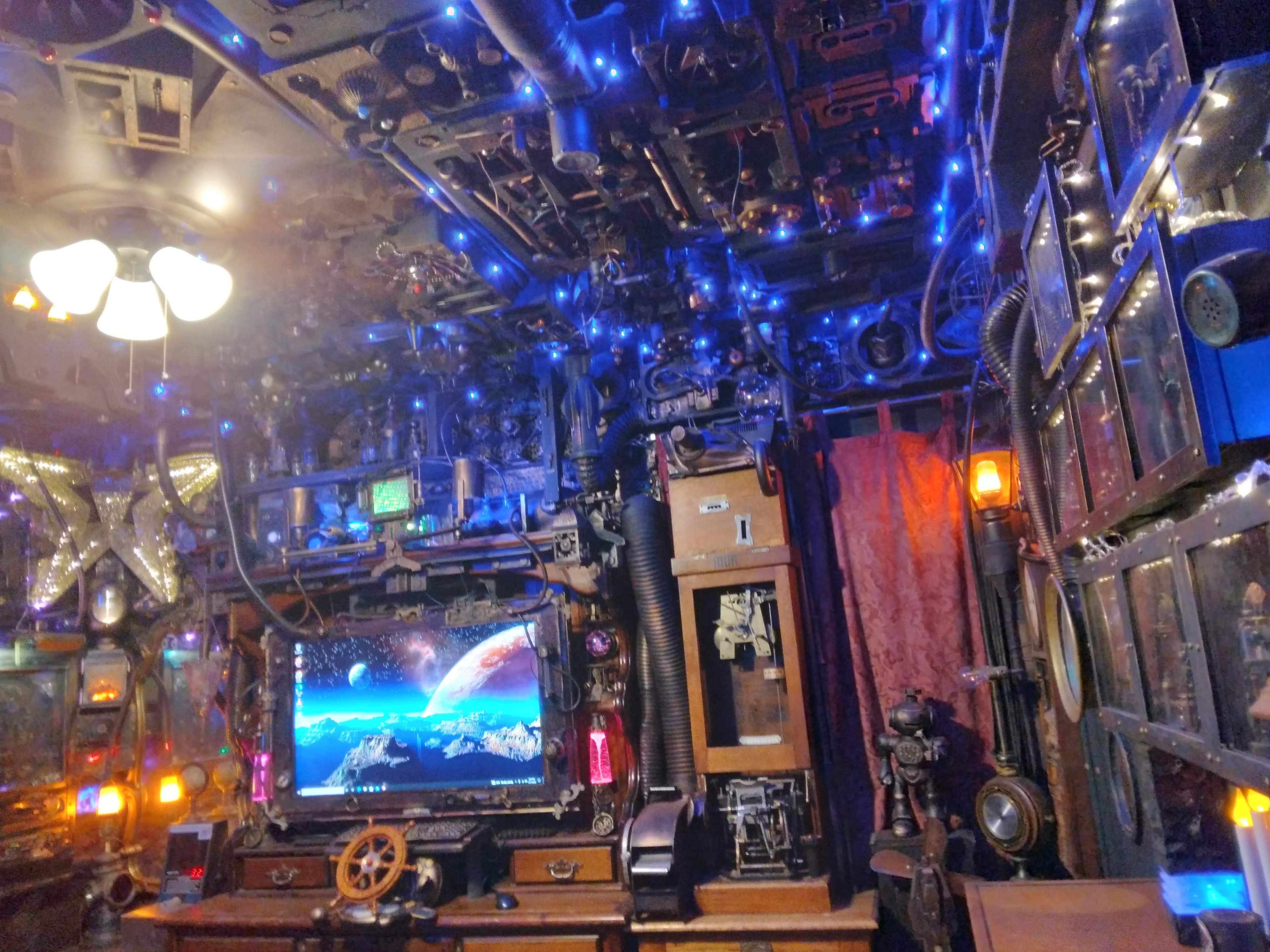 The image depicts a cluttered room filled with various mechanical objects, vintage decorations, and blue lights, with a large screen displaying a space scene.
