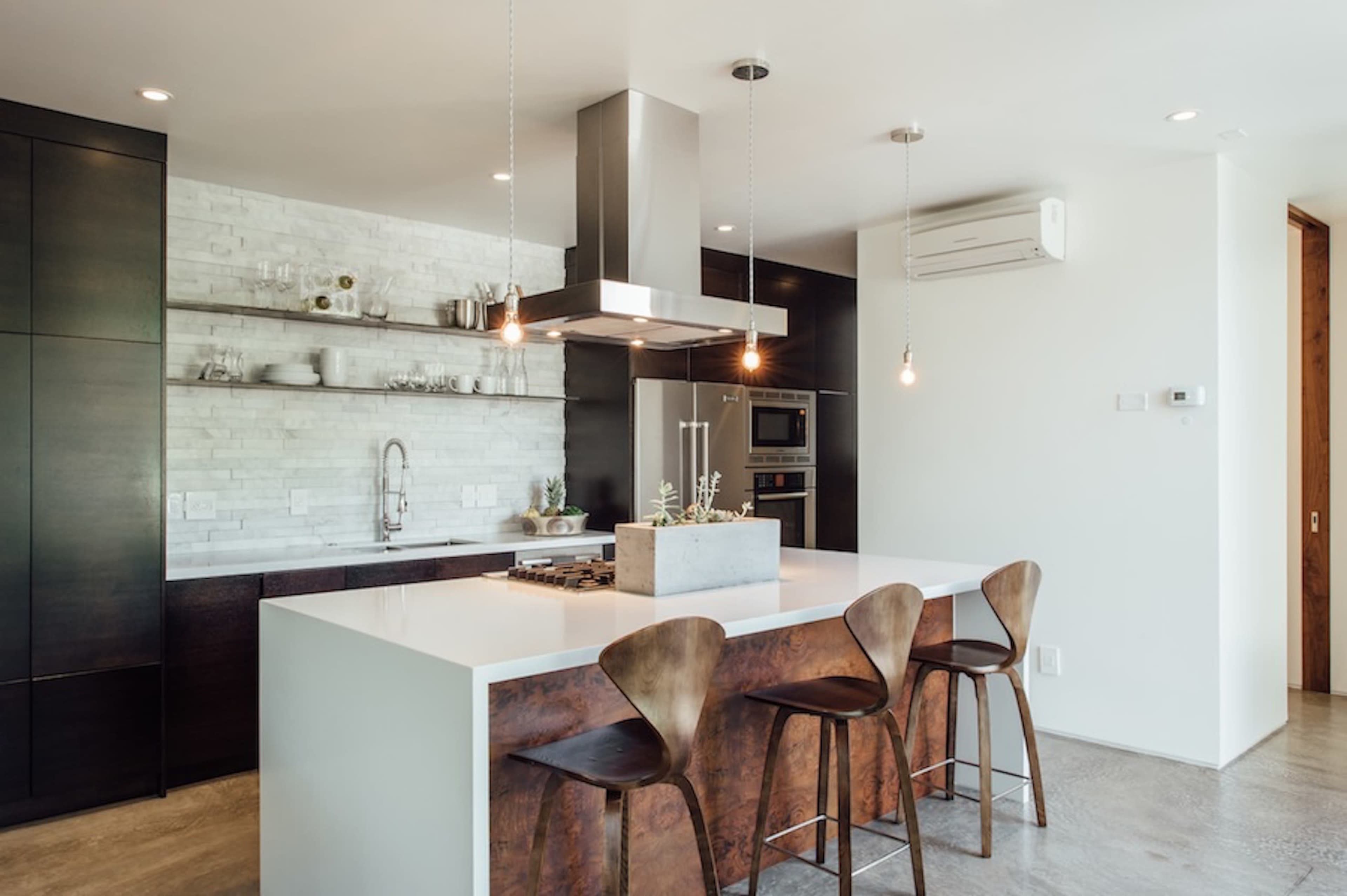 The image shows a modern kitchen featuring a central island with four wooden stools, a stainless steel range hood, and a minimalist design.