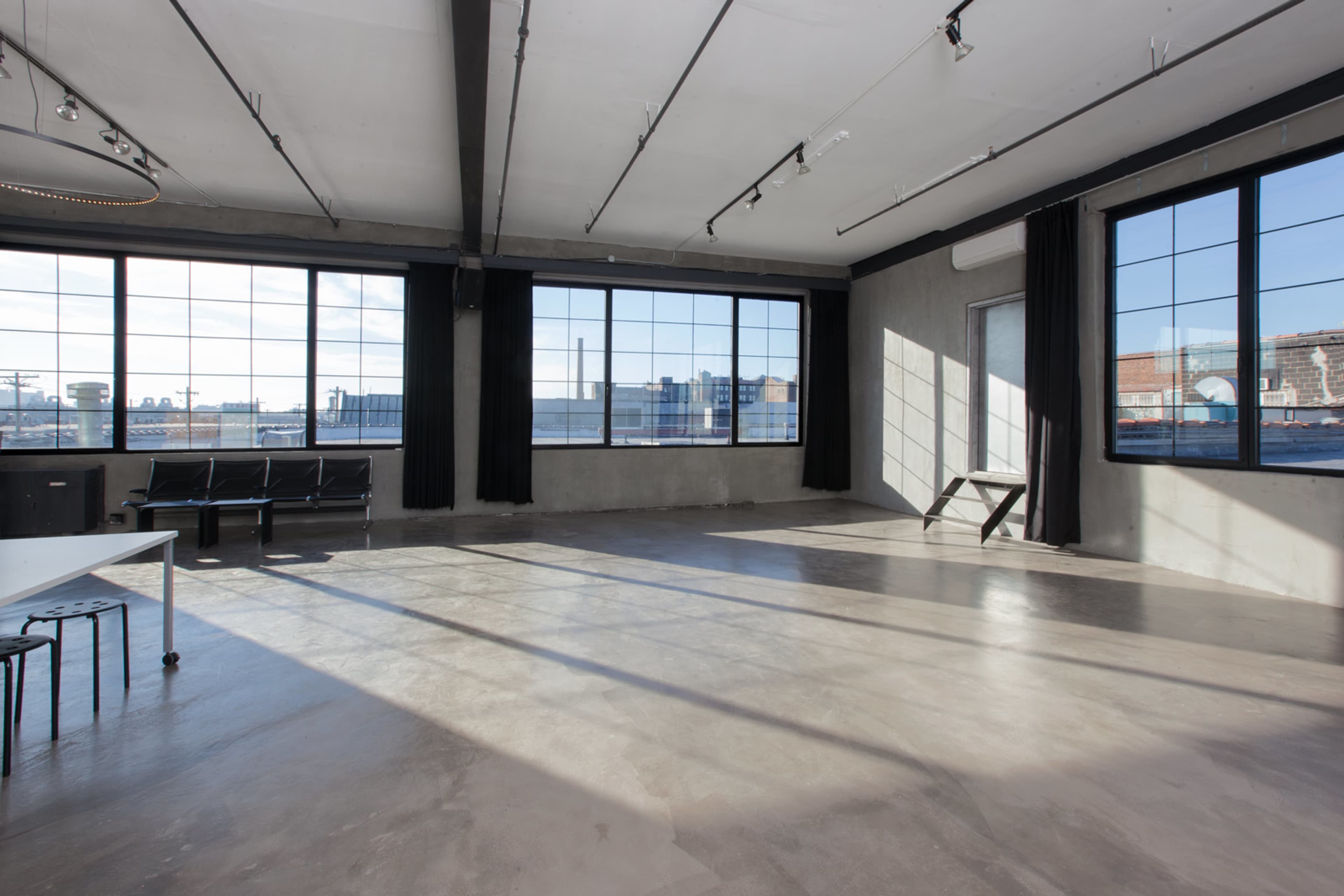 The image shows an empty industrial-style room with large windows, concrete flooring, and minimal furnishings.
