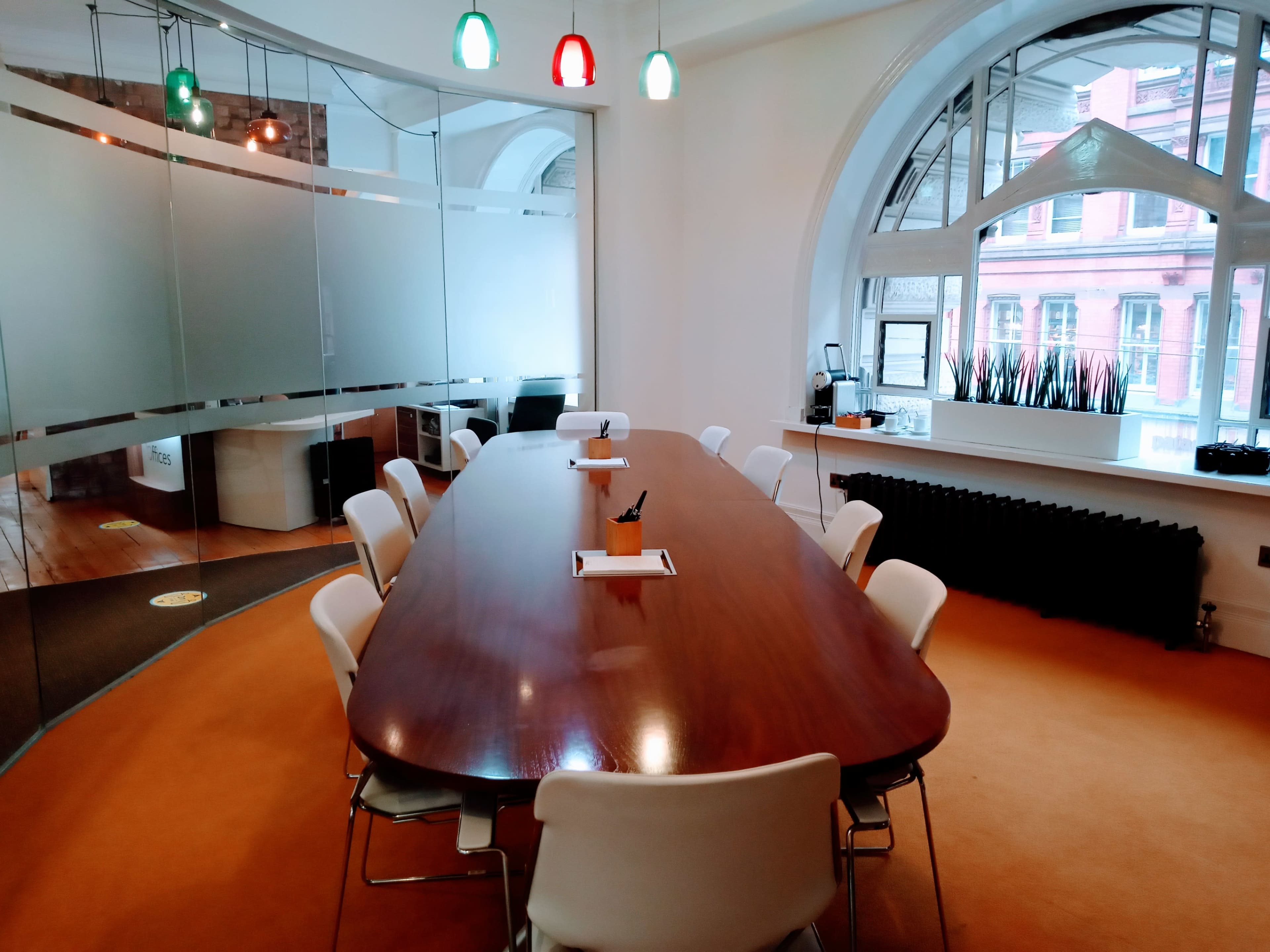 A long wooden conference table is surrounded by white chairs in a well-lit meeting room with colorful overhead lights and a glass partition.