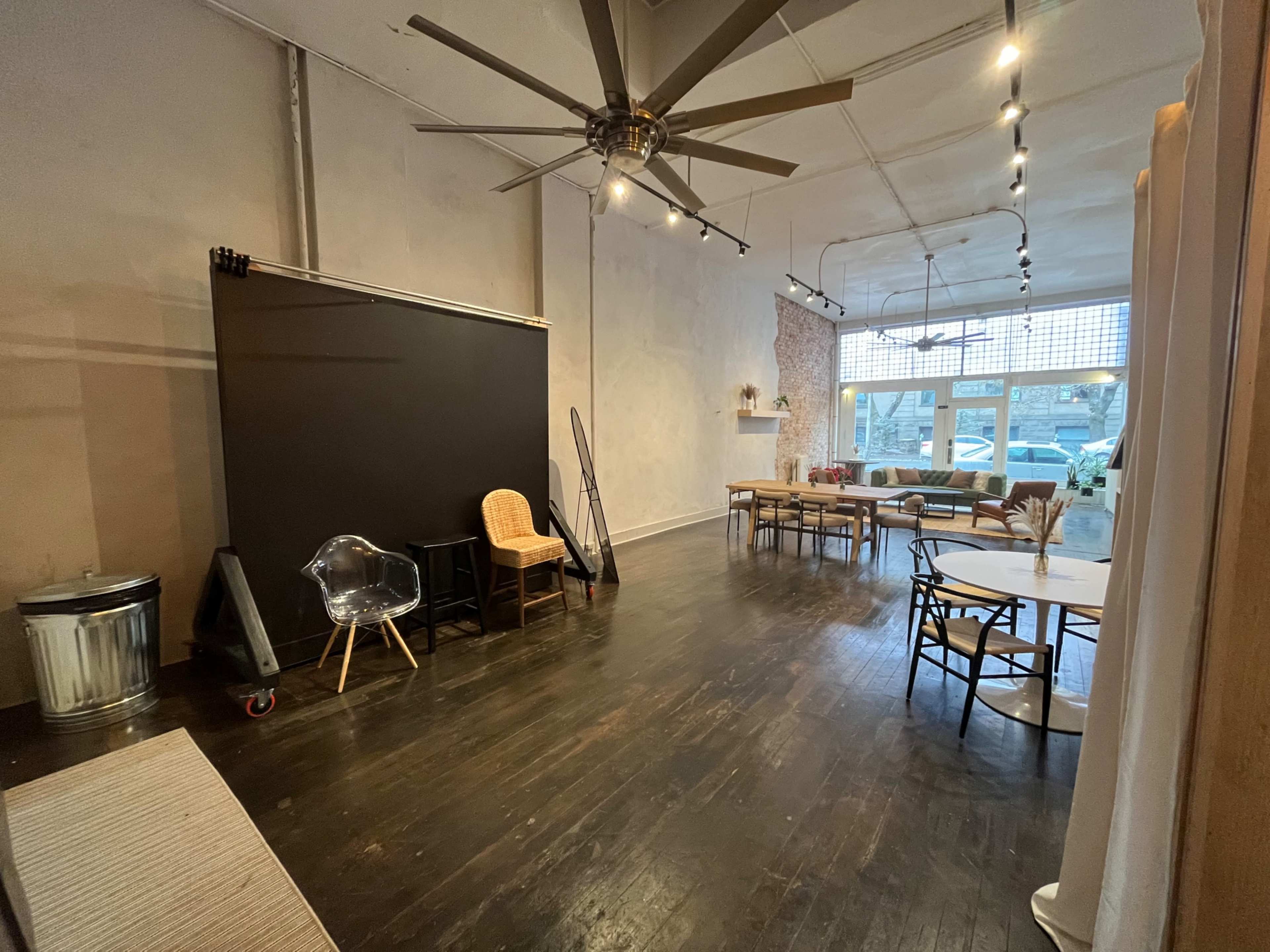 An empty interior space with a large ceiling fan, various seating options, a black backdrop, and a row of windows along one wall.