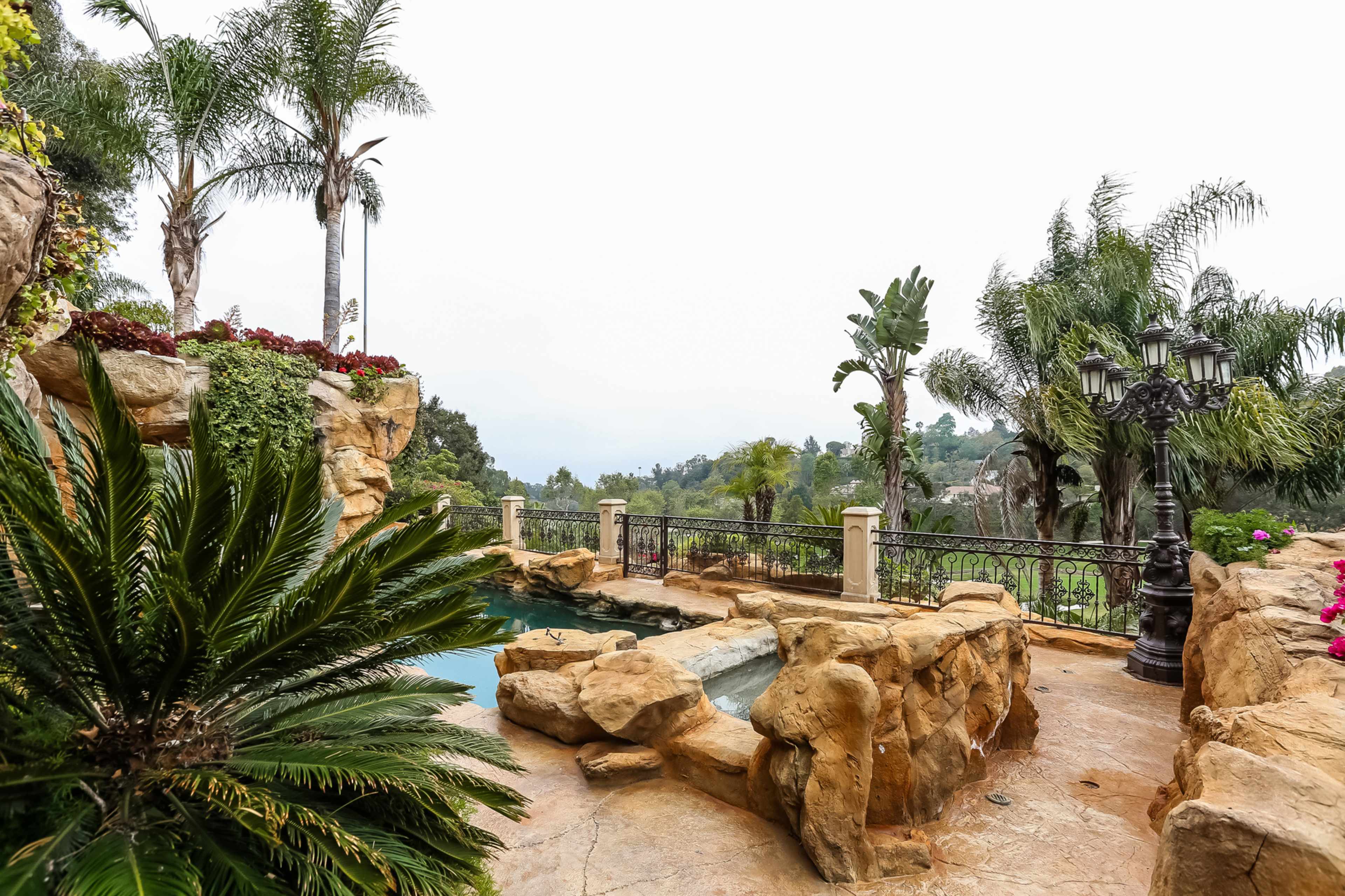 A landscaped outdoor area with a stone patio, lush greenery, and a swimming pool overlooking a distant view.