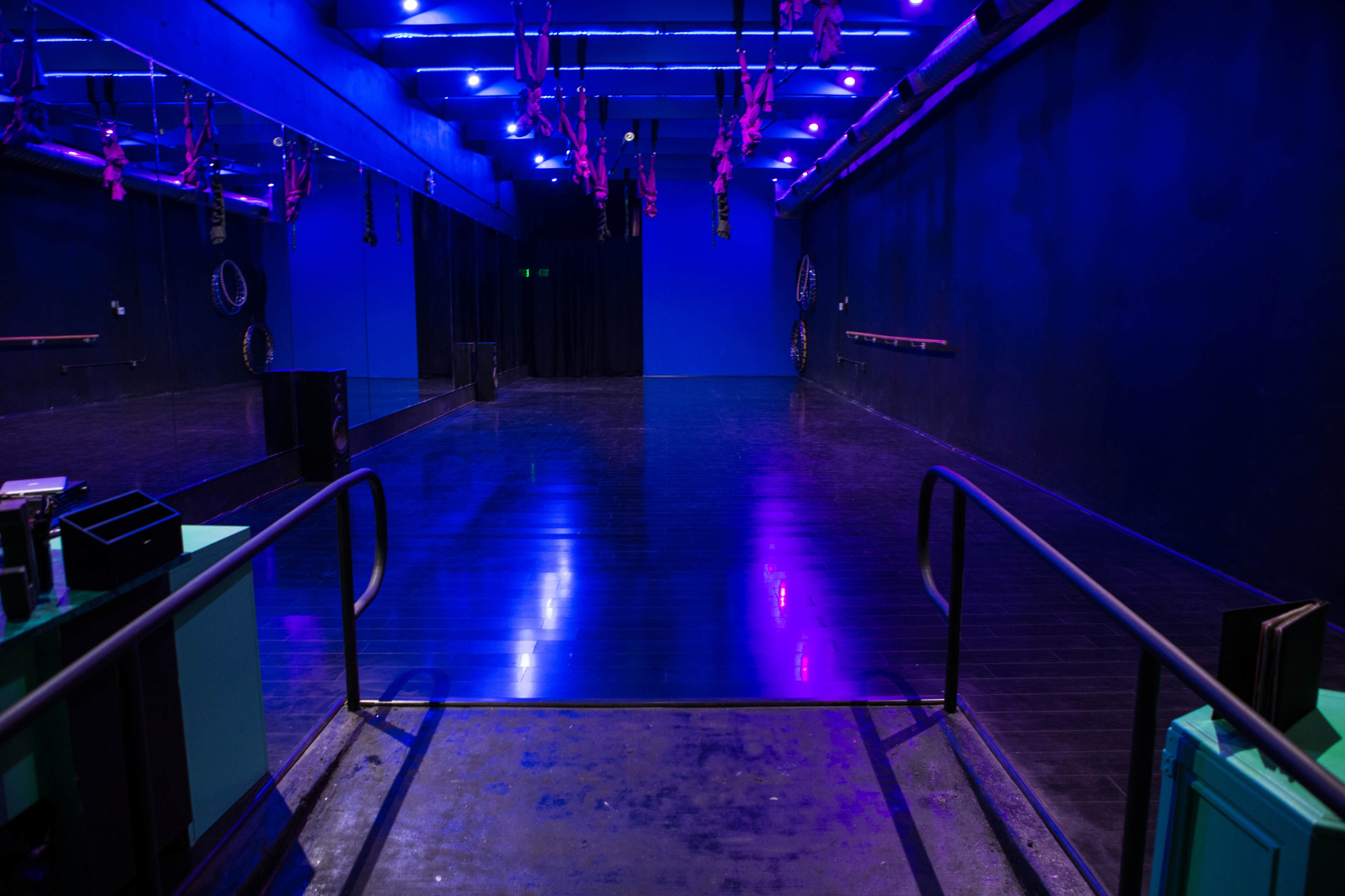 A spacious dance studio features a polished floor, vibrant blue walls, and overhead purple lighting.