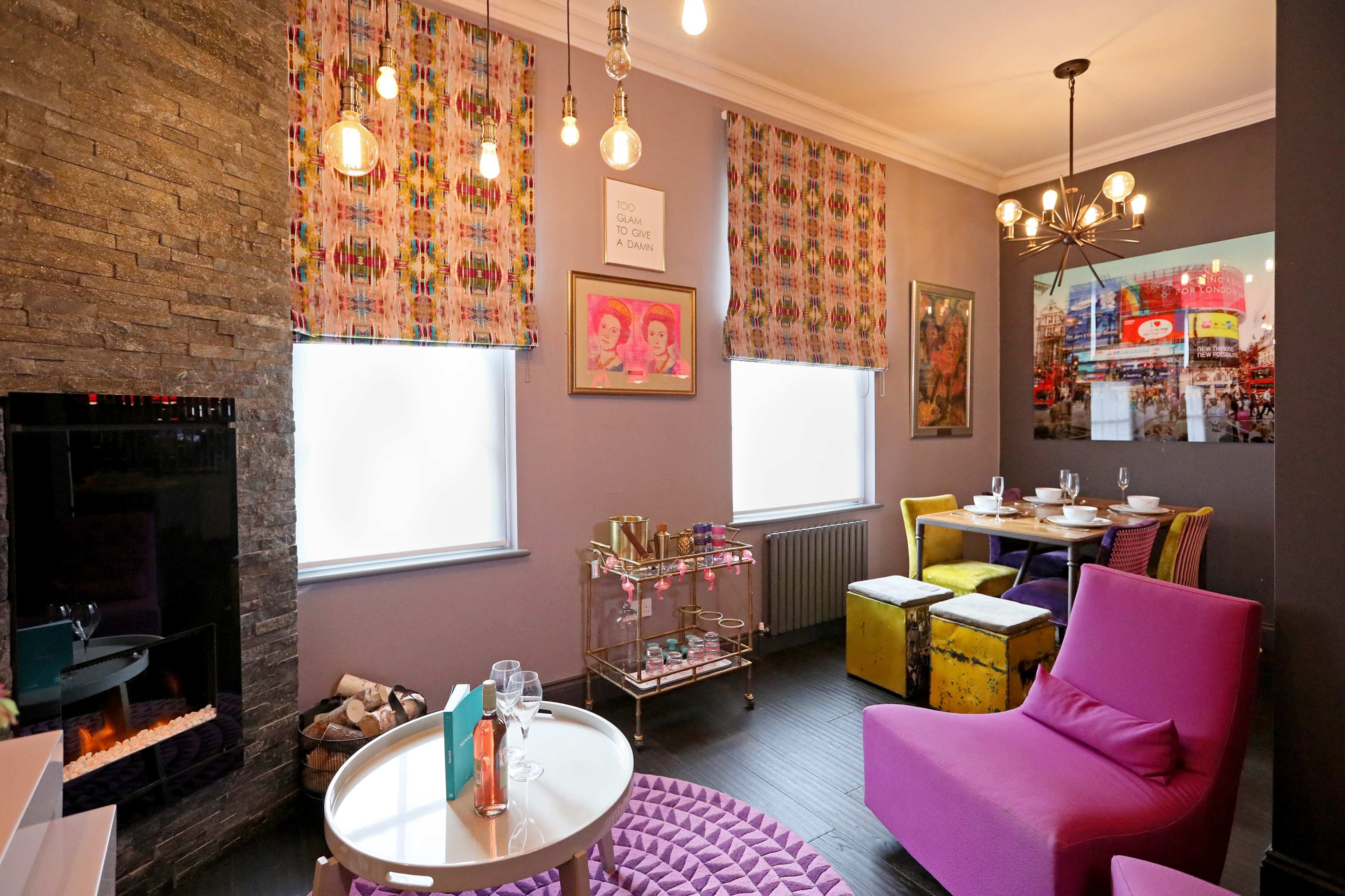 A modern dining area with a stone wall, colorful window treatments, and a mix of seating options, including a pink armchair and a dining table set for guests.