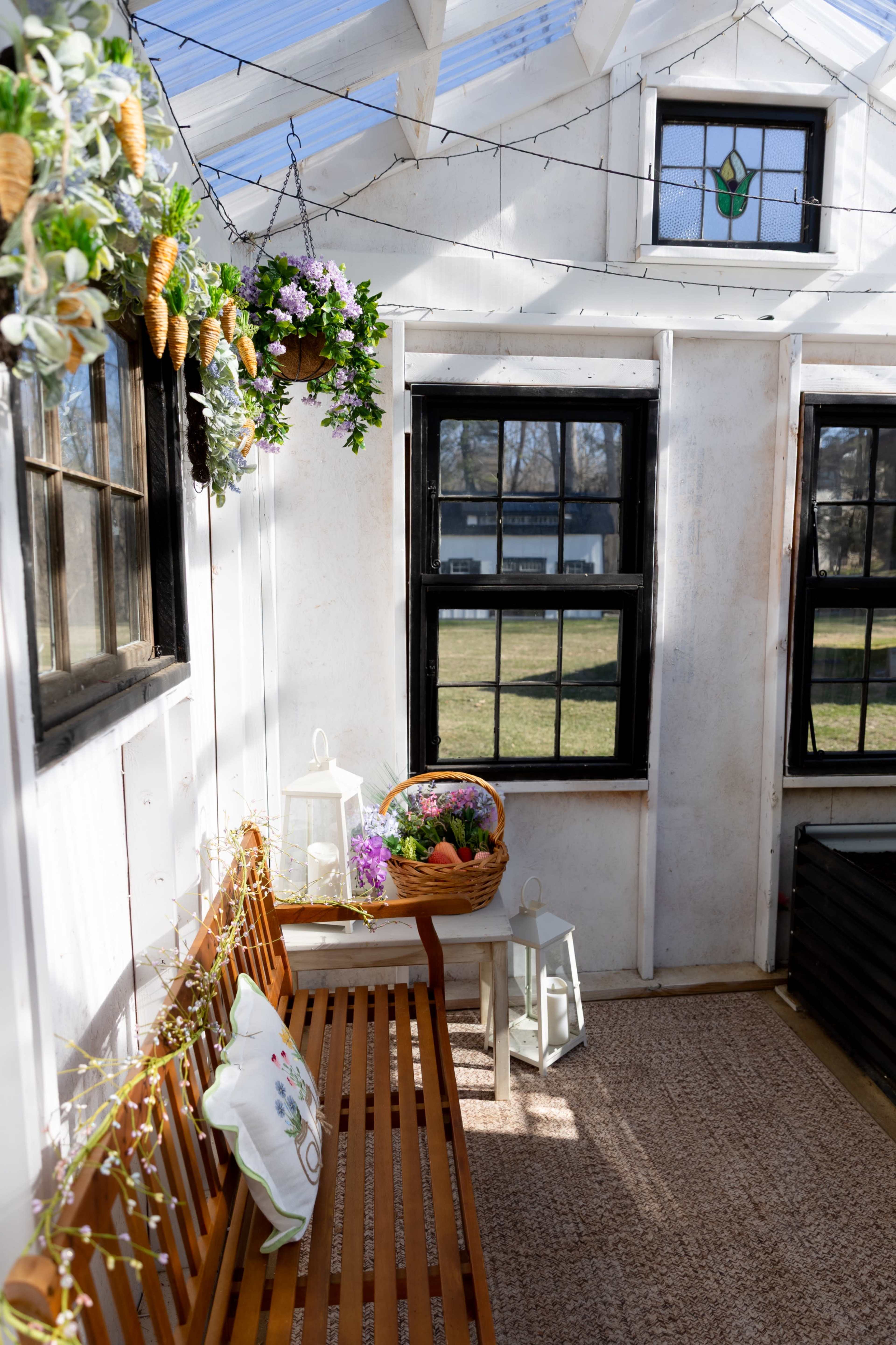 The image shows a sunlit, white-walled interior of a greenhouse featuring a wooden bench, potted plants, and decorative hanging elements near large windows.