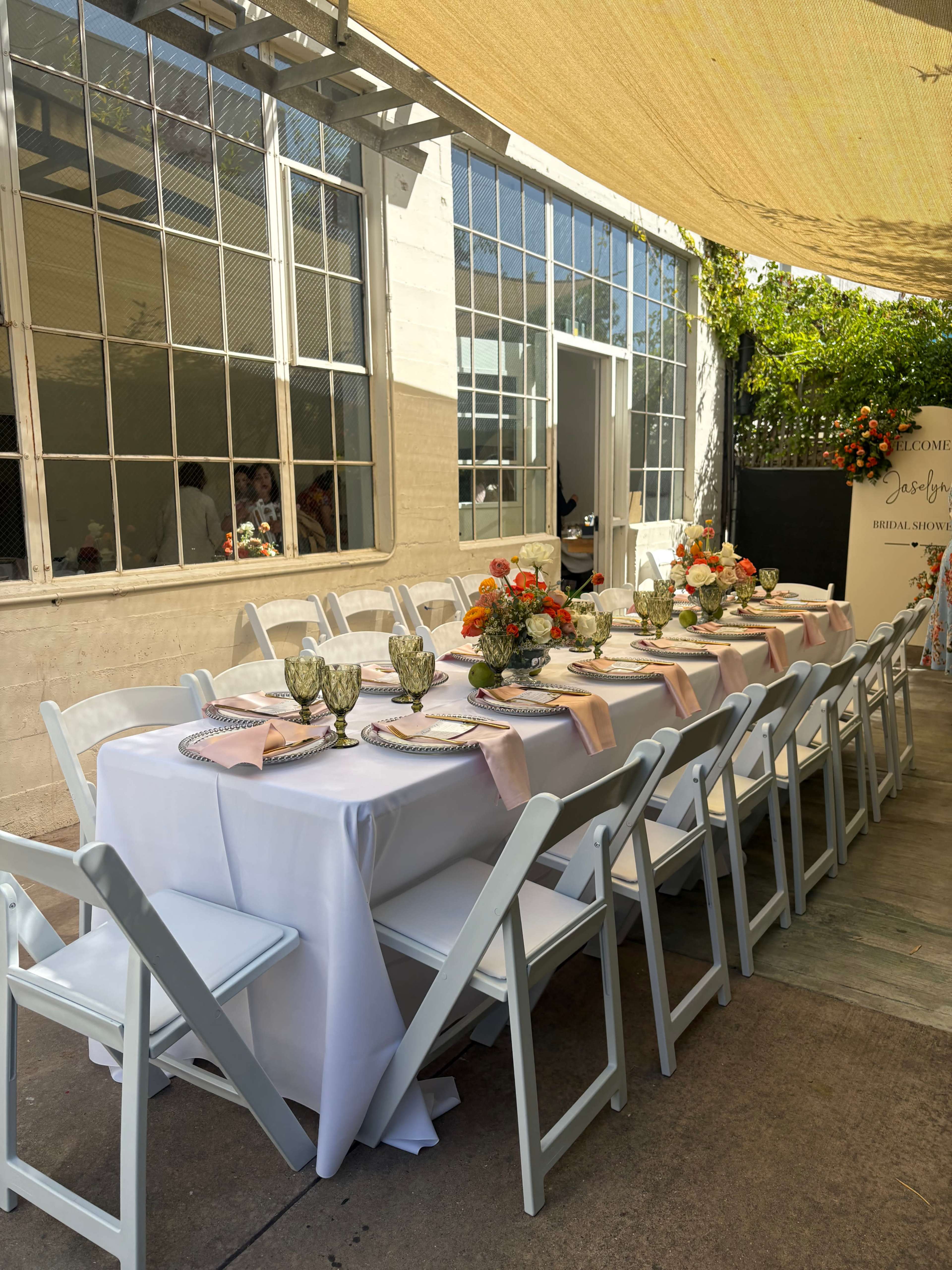 A long dining table set for a gathering outdoors features white tablecloths, floral centerpieces, and chairs lined along each side.