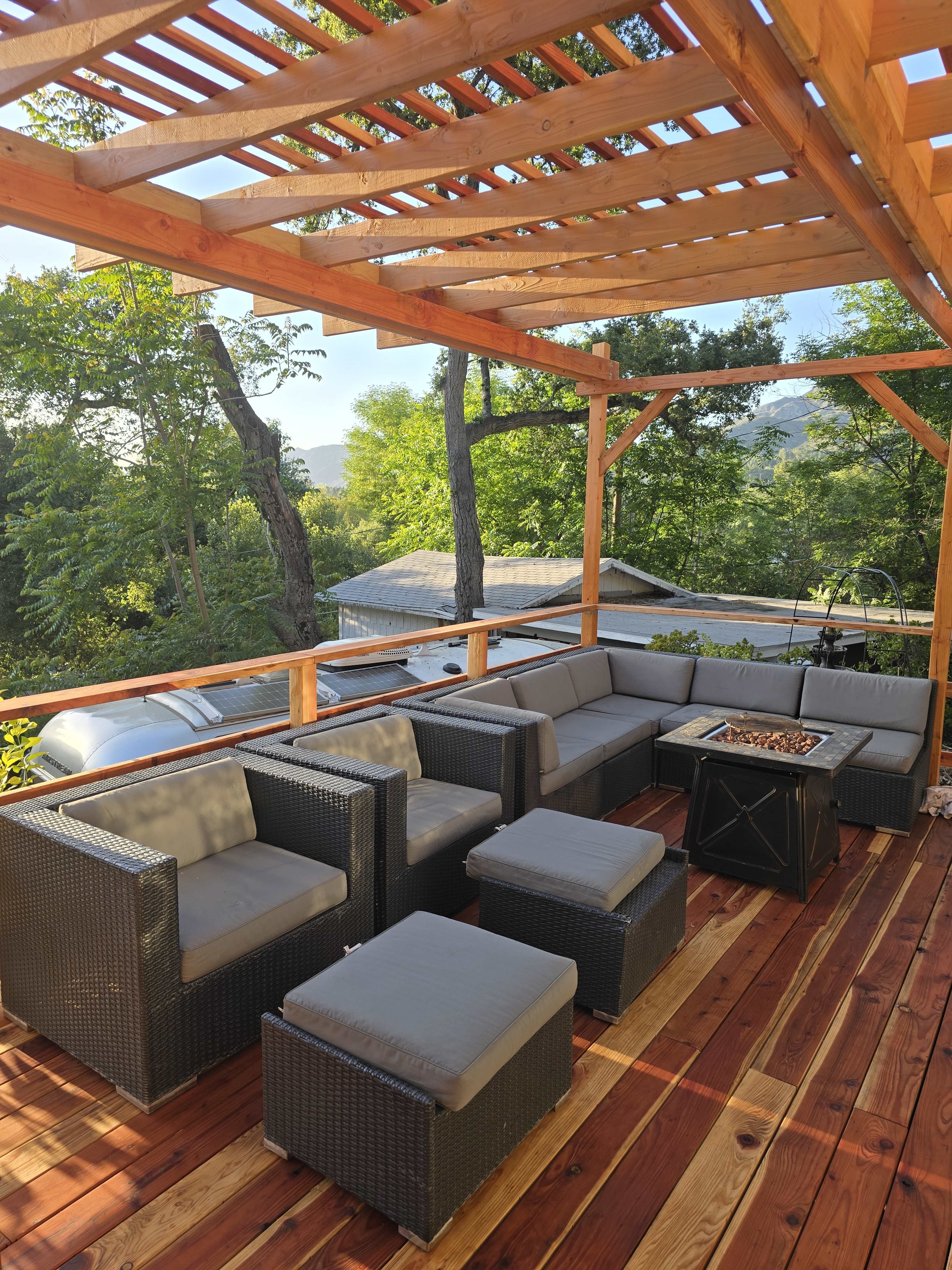 The image shows a wooden patio with a seating area comprised of black wicker furniture and a central fire pit, surrounded by trees.