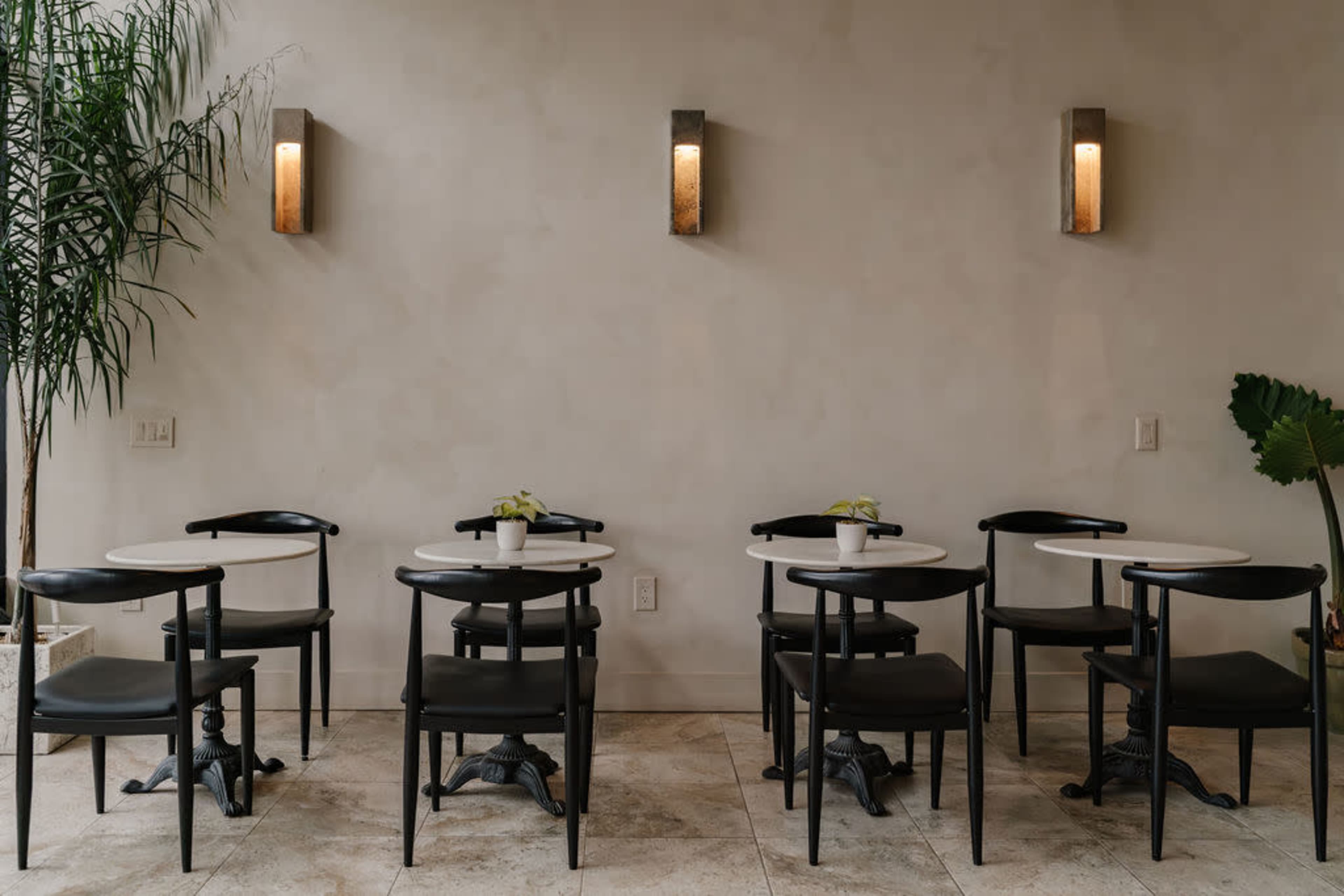 The image shows a minimalistic café setting with several small round tables and black chairs arranged against a neutral wall, illuminated by wall sconces.