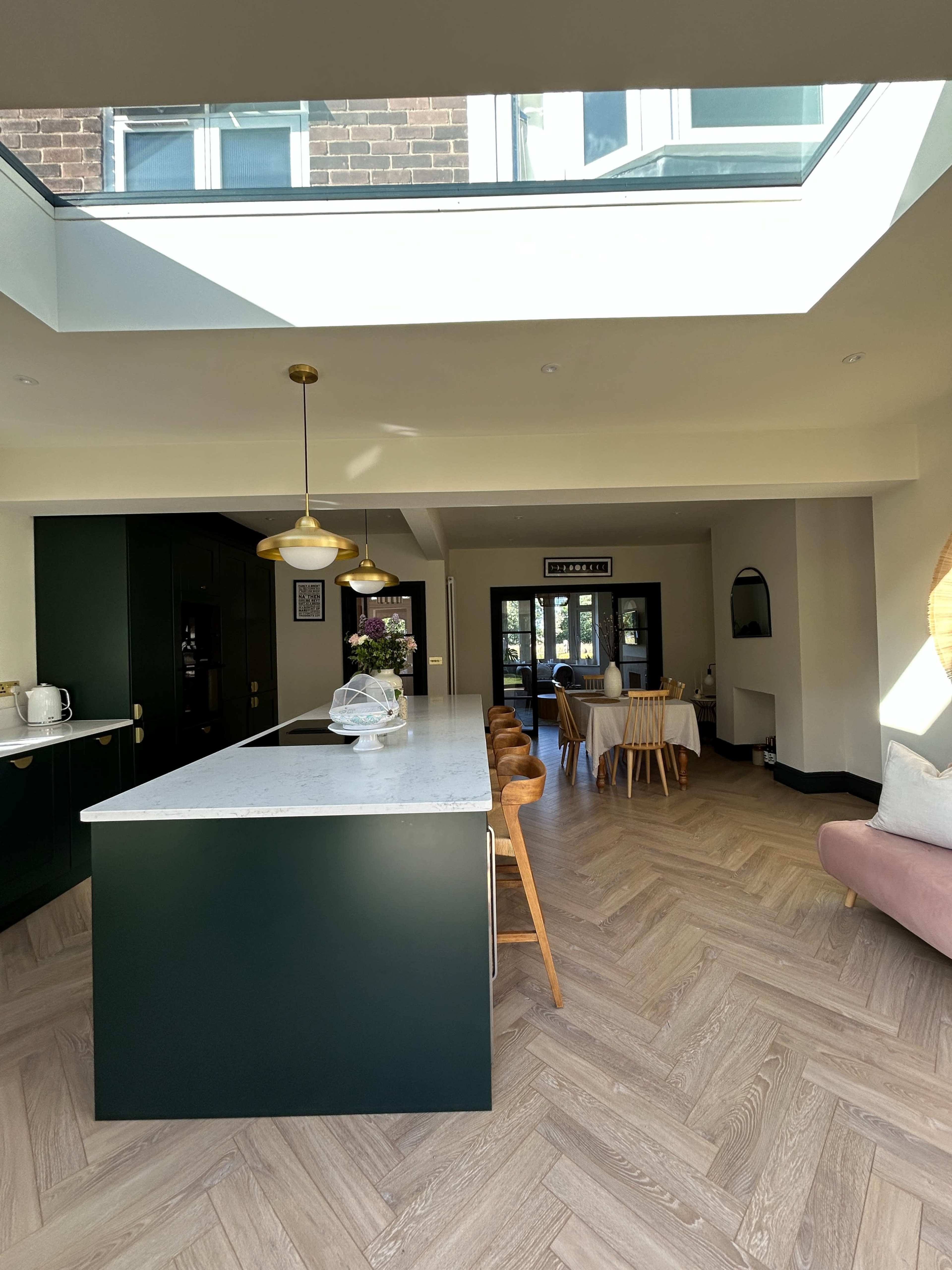 The image shows a modern kitchen with a large island, wooden dining chairs, and a skylight allowing natural light to enter the space.