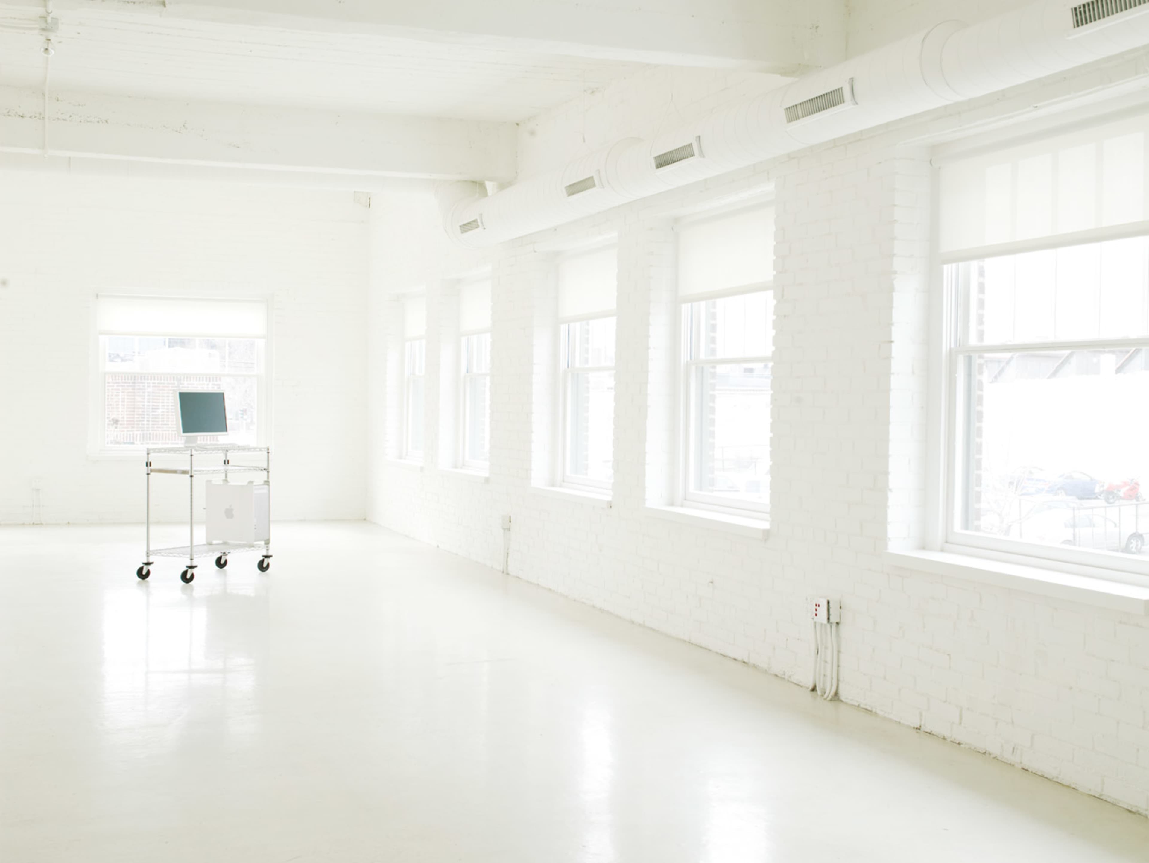 The image shows a bright, minimalistic white room with large windows and a laptop on a cart in the center.