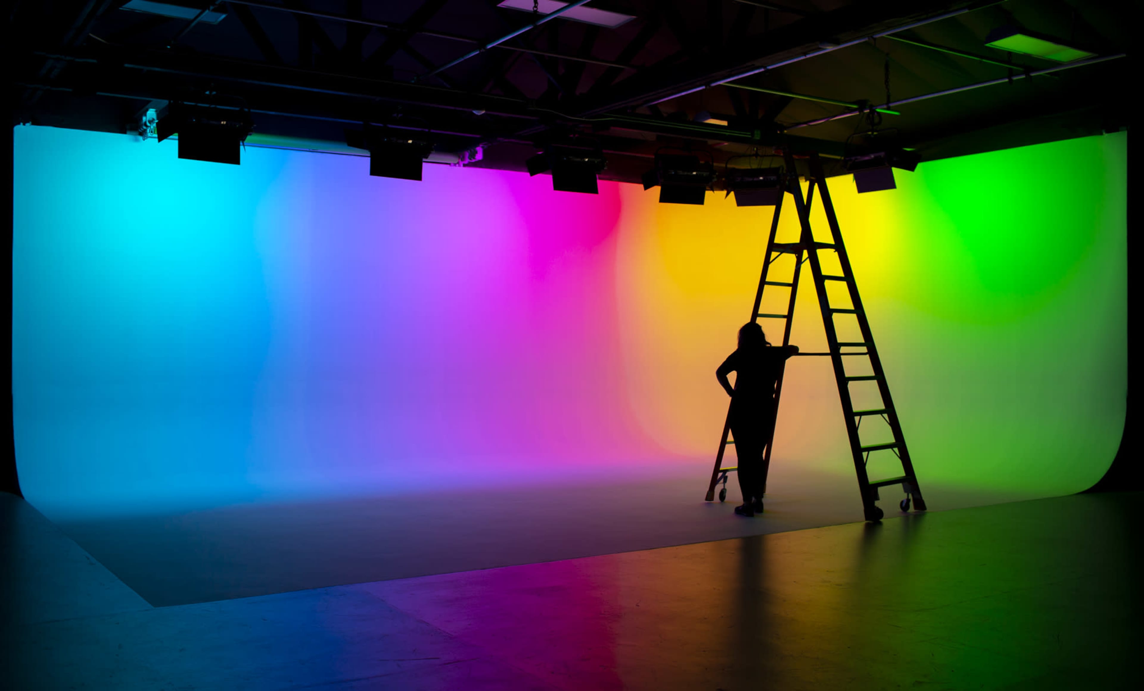 A person stands on a ladder in front of a brightly colored gradient backdrop in a studio.