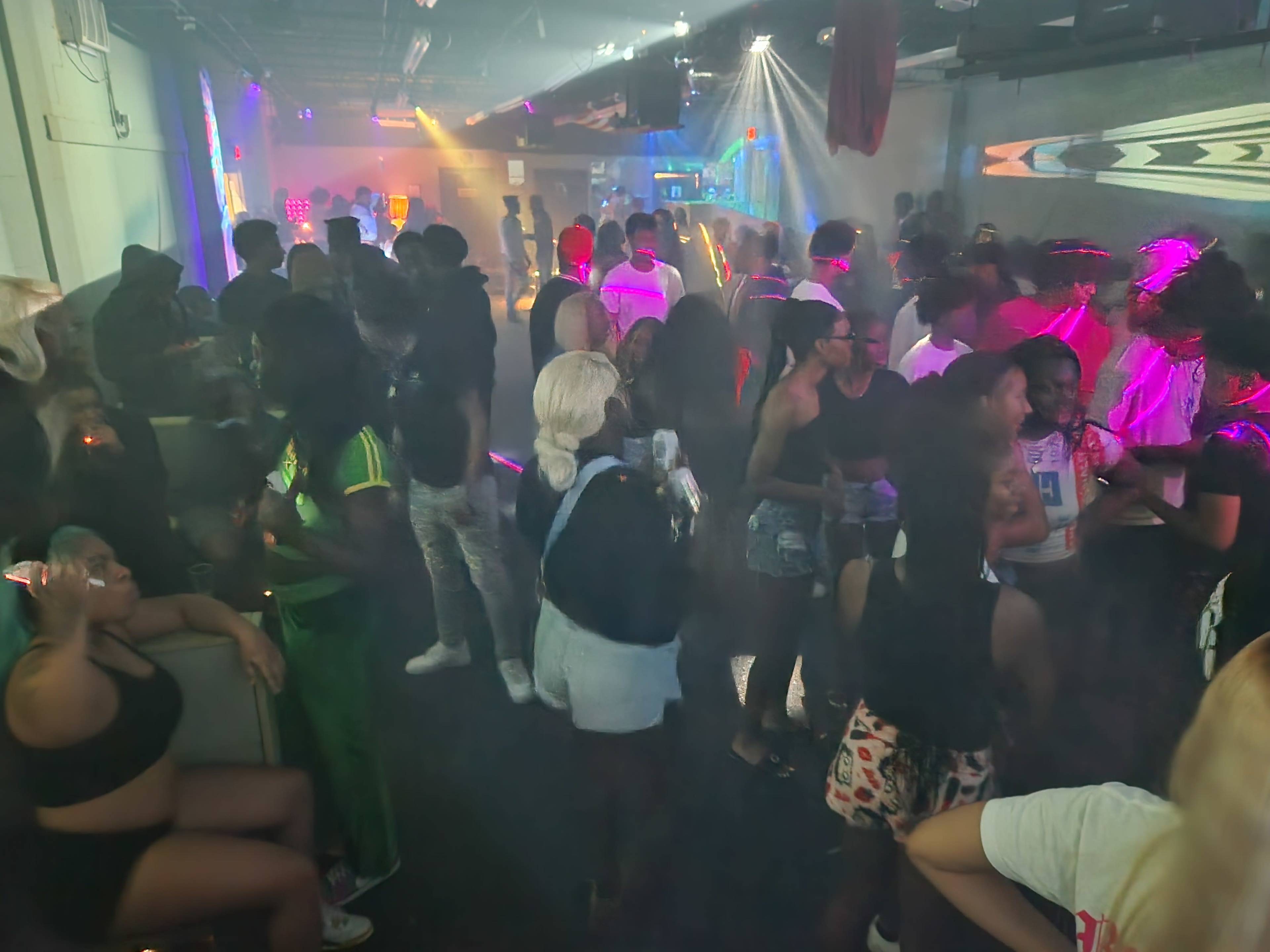 The image shows a crowded room filled with people socializing in a dimly lit environment enhanced by colorful lights and smoke effects.