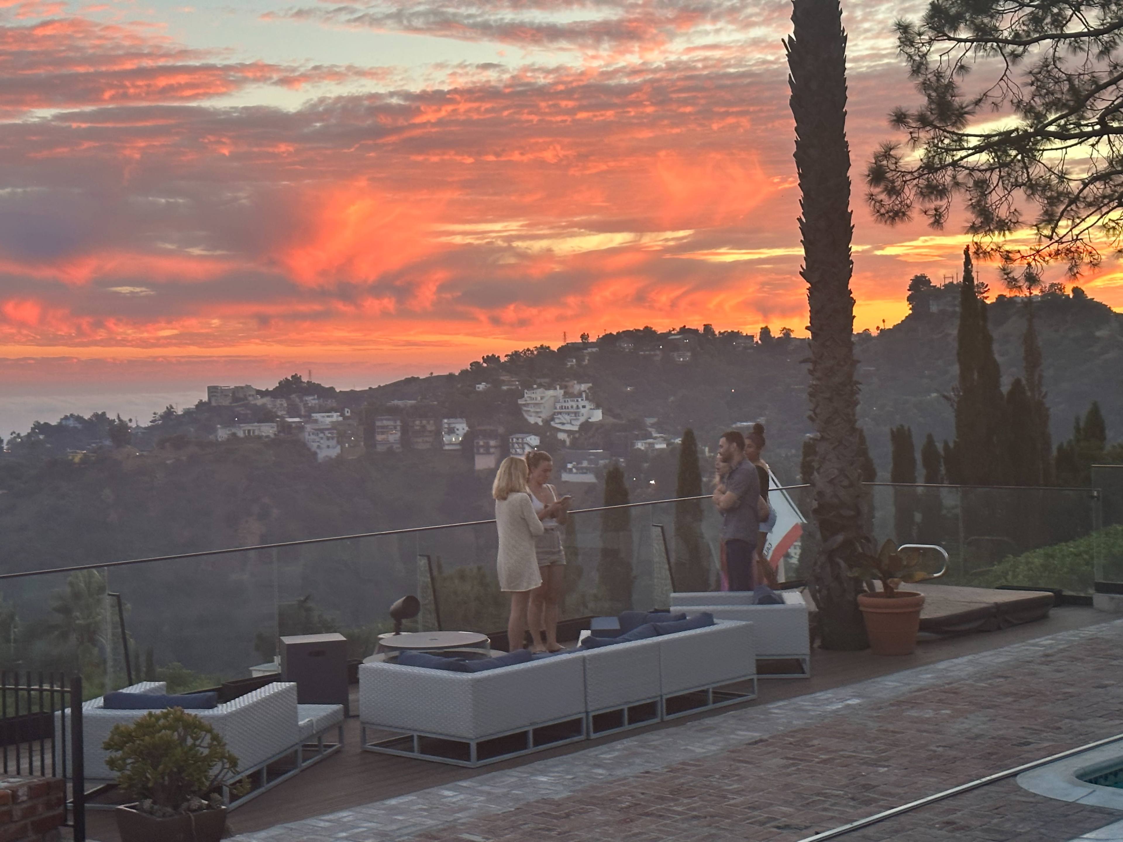 A group of people stands on a deck overlooking a hillside at sunset, with colorful clouds illuminating the sky.