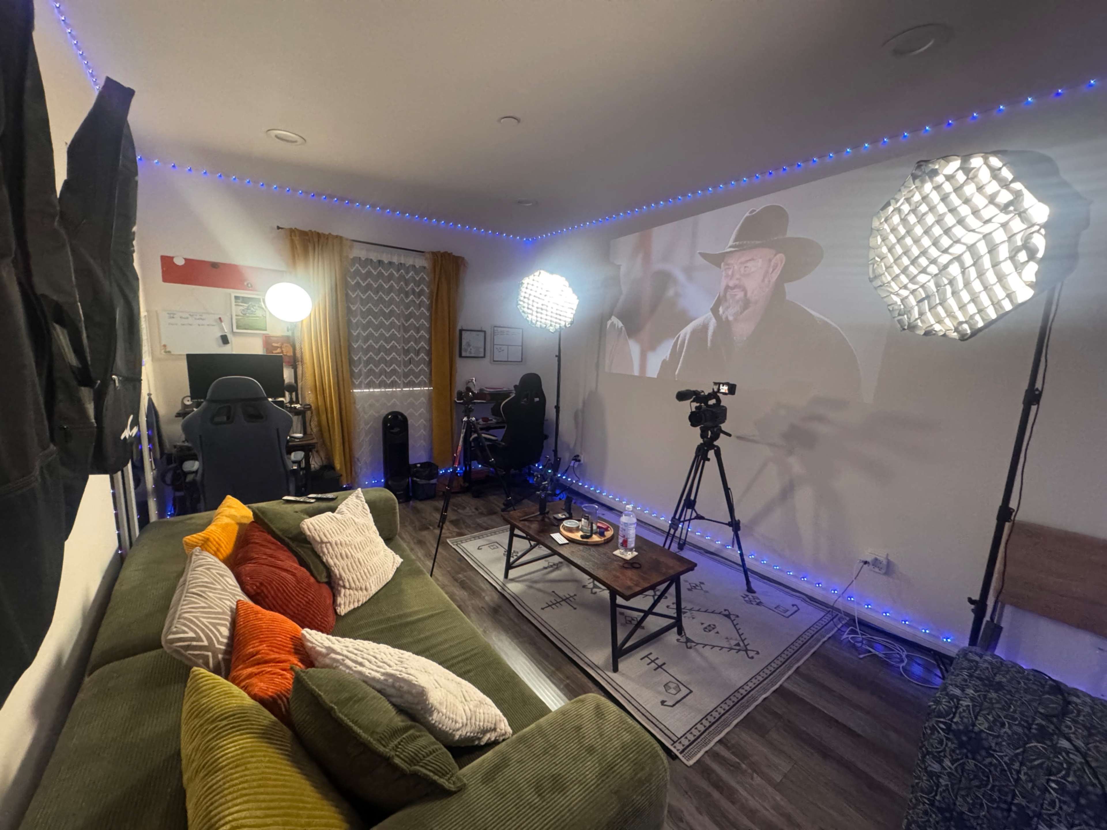 The image shows a cozy room with a green sectional couch adorned with colorful pillows, a projector displaying a film on the wall, and two softbox lights set up for video recording.