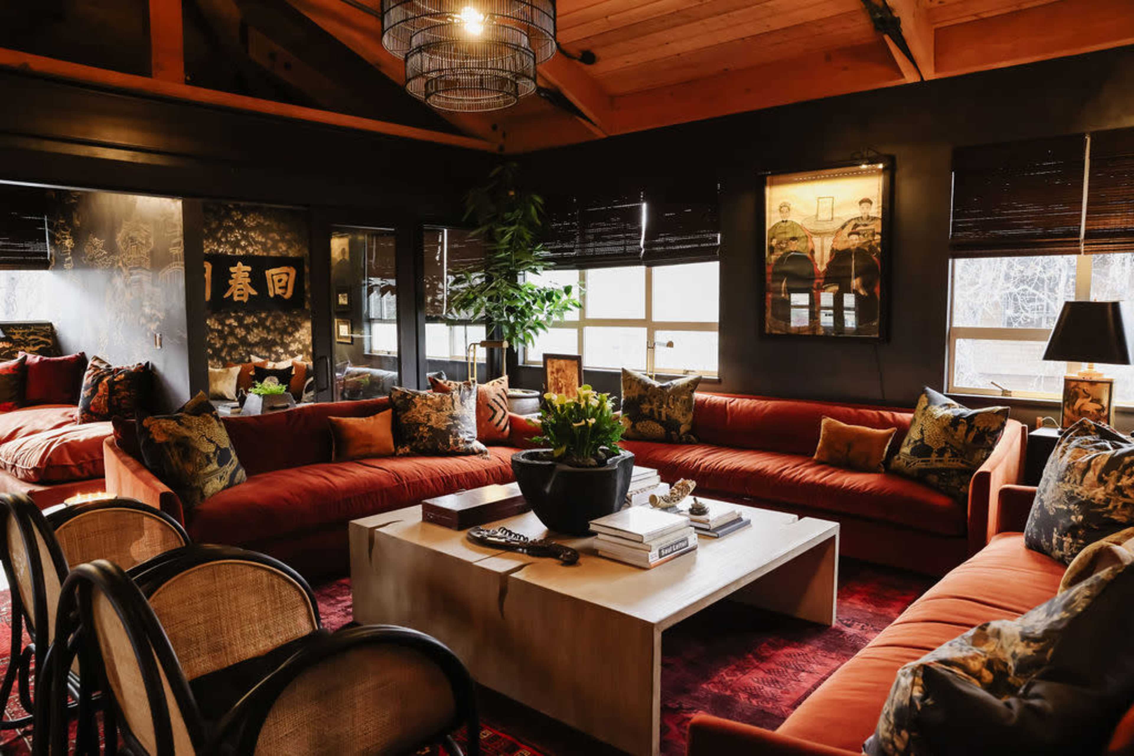 A spacious living room with large, orange sofas, wooden beams, and a central coffee table surrounded by decorative pillows and plants.