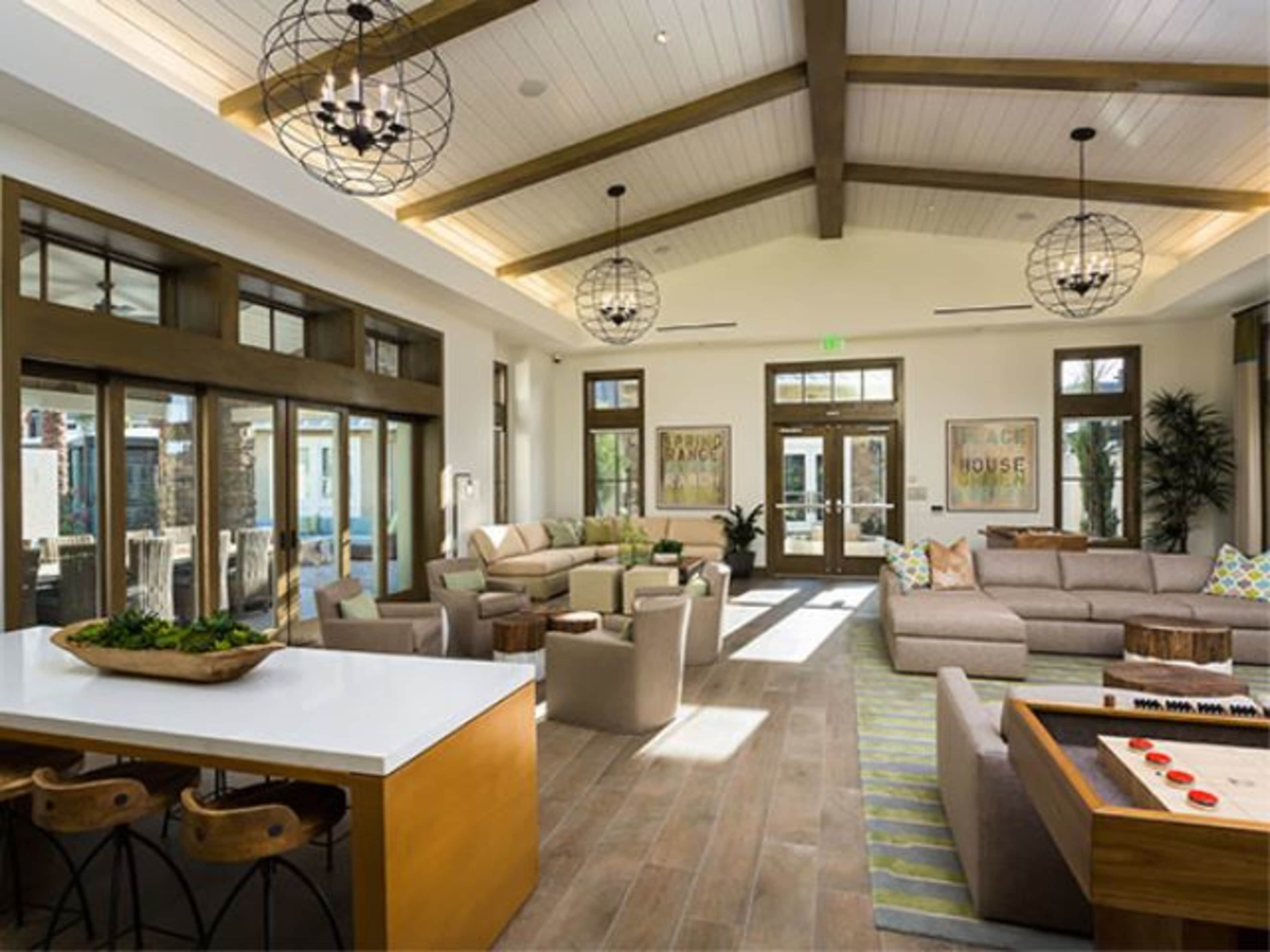 A spacious living room with a high wooden ceiling, featuring comfortable furniture, large windows, and a kitchen area.
