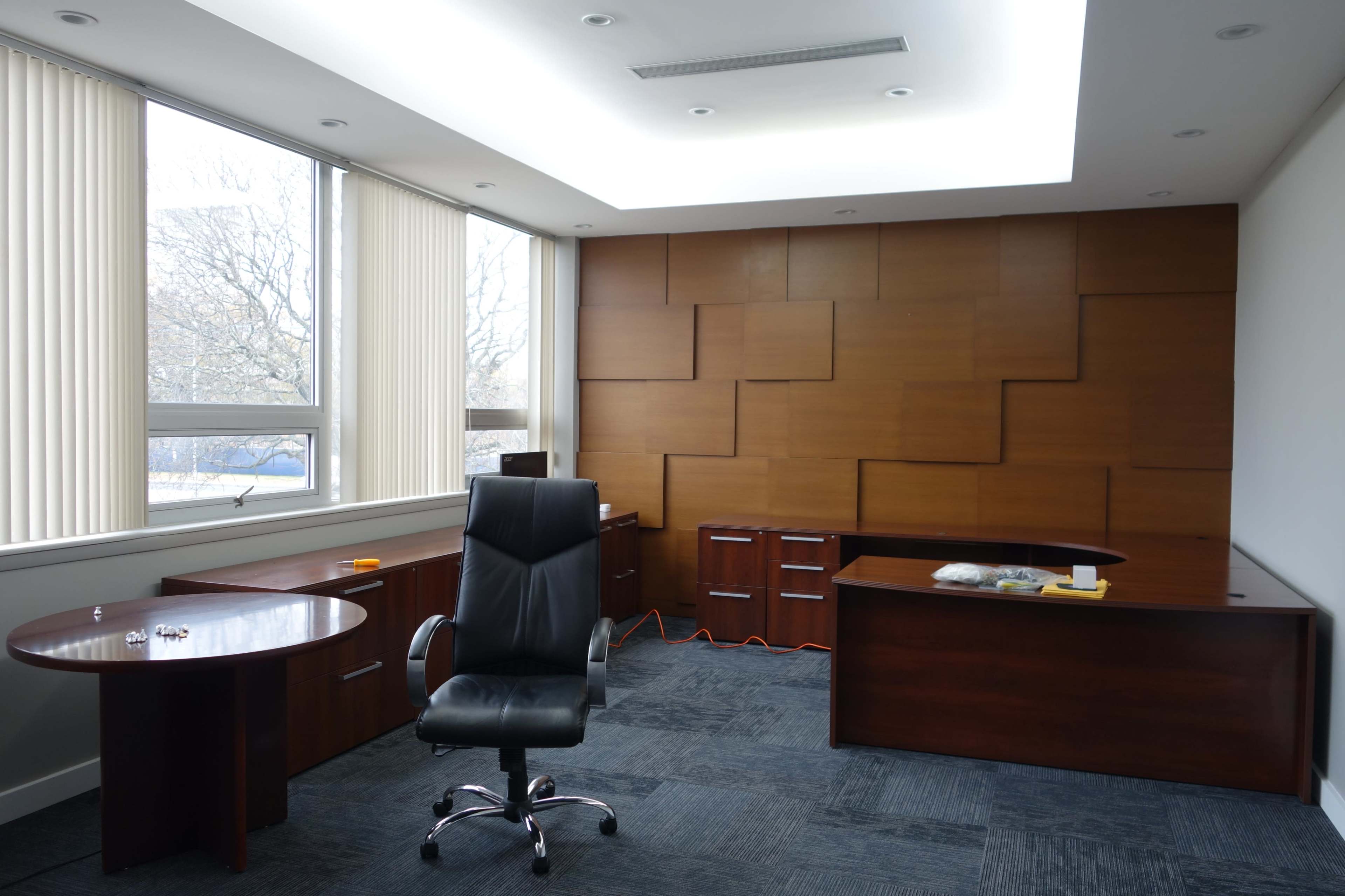 A modern office interior with a black ergonomic chair, a round table, and wooden cabinets against a wall featuring geometric paneling.