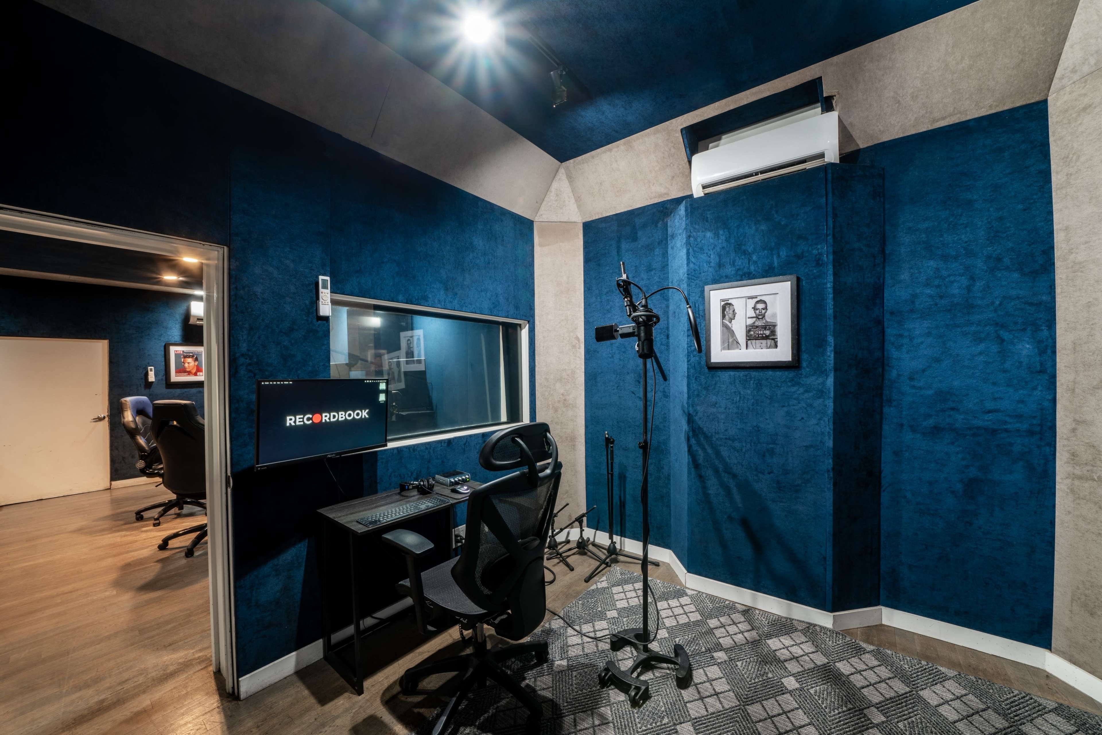 The image shows a recording studio with blue walls, a soundproof booth, a desk with a computer, and a microphone setup.