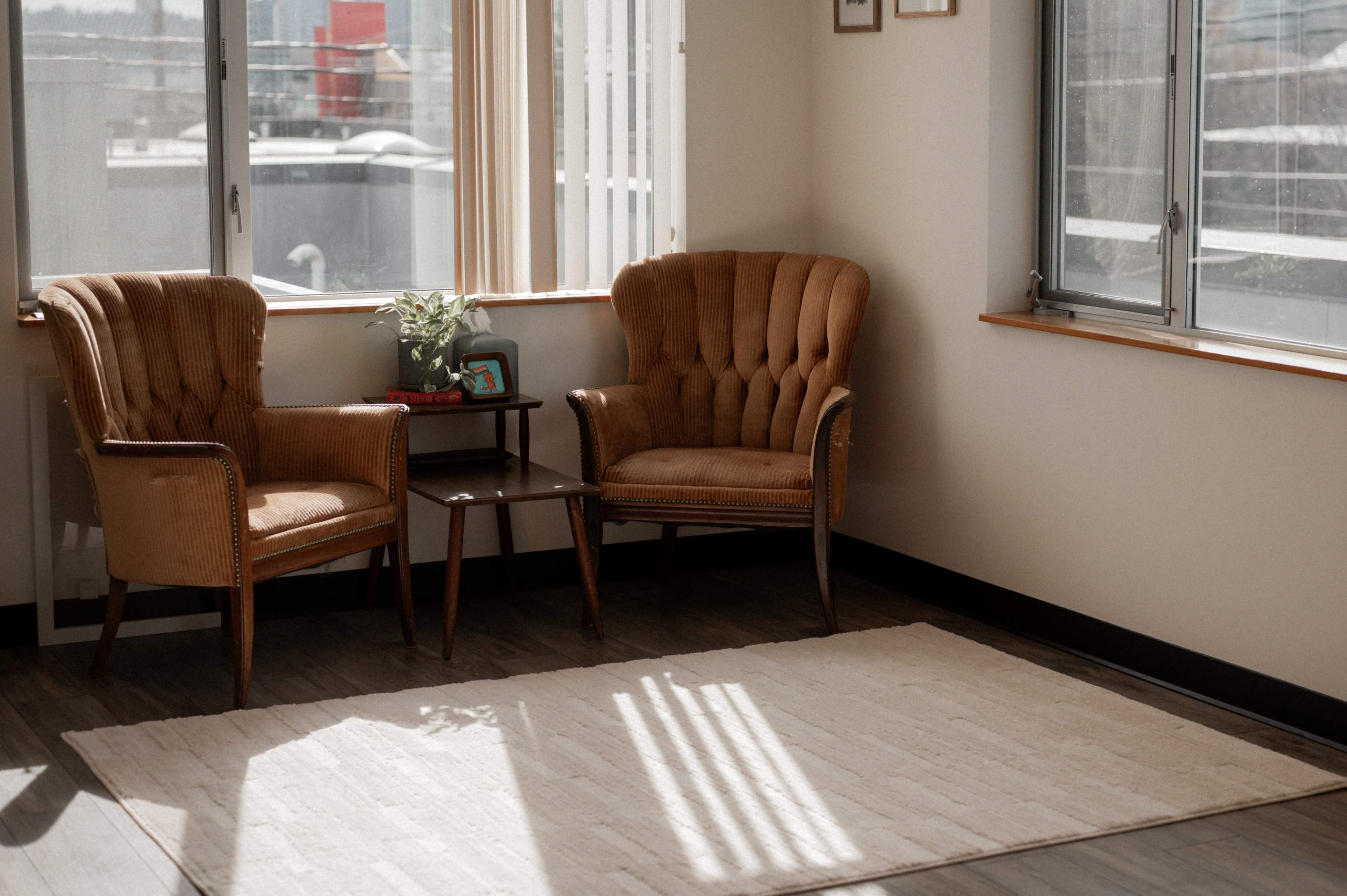 The image shows a bright corner of a room with two vintage armchairs and a small table between them, next to a window.
