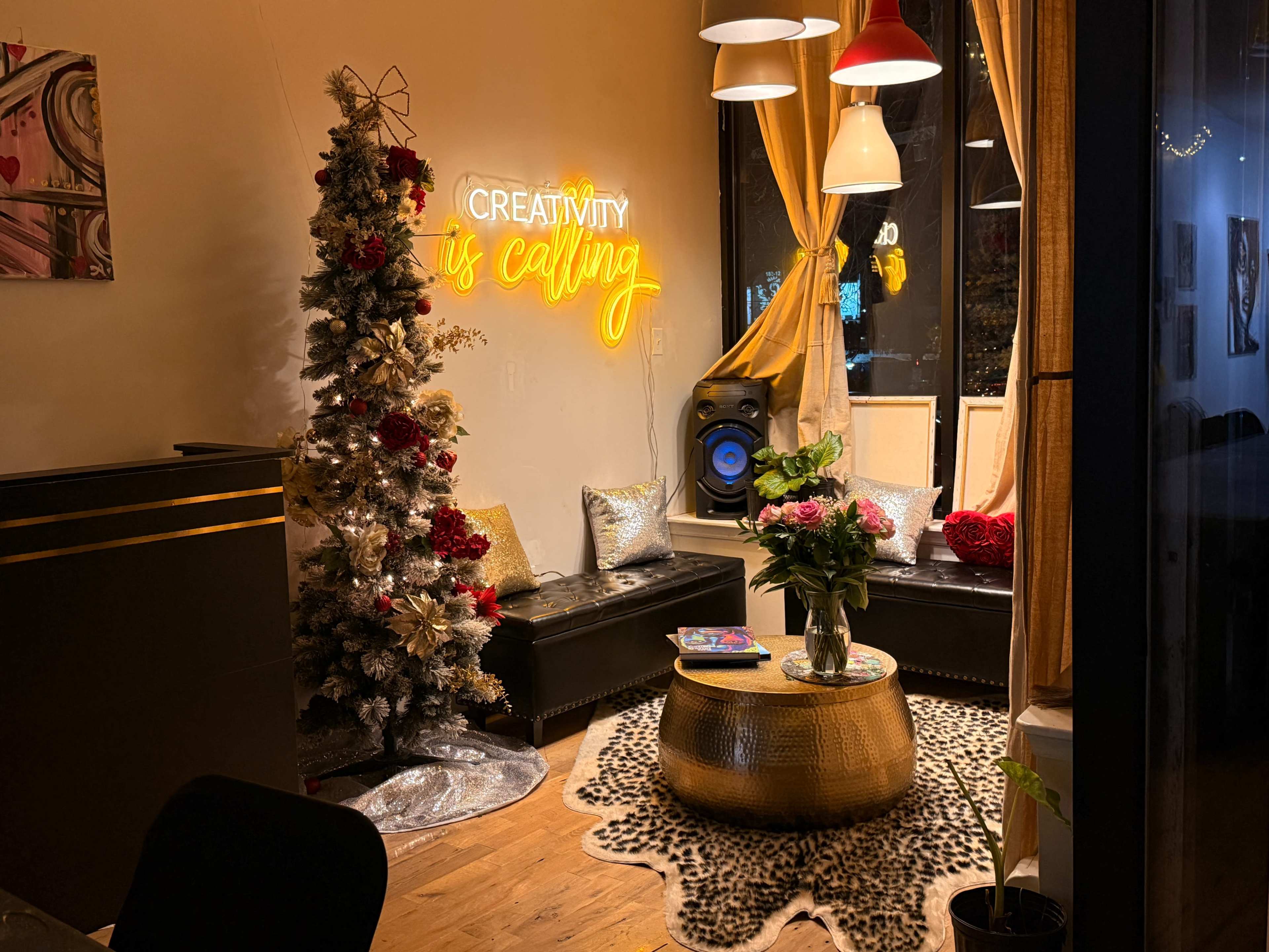 The image shows a cozy interior space with a decorative Christmas tree, a neon sign that reads "Creativity is calling," and a round table adorned with flowers.