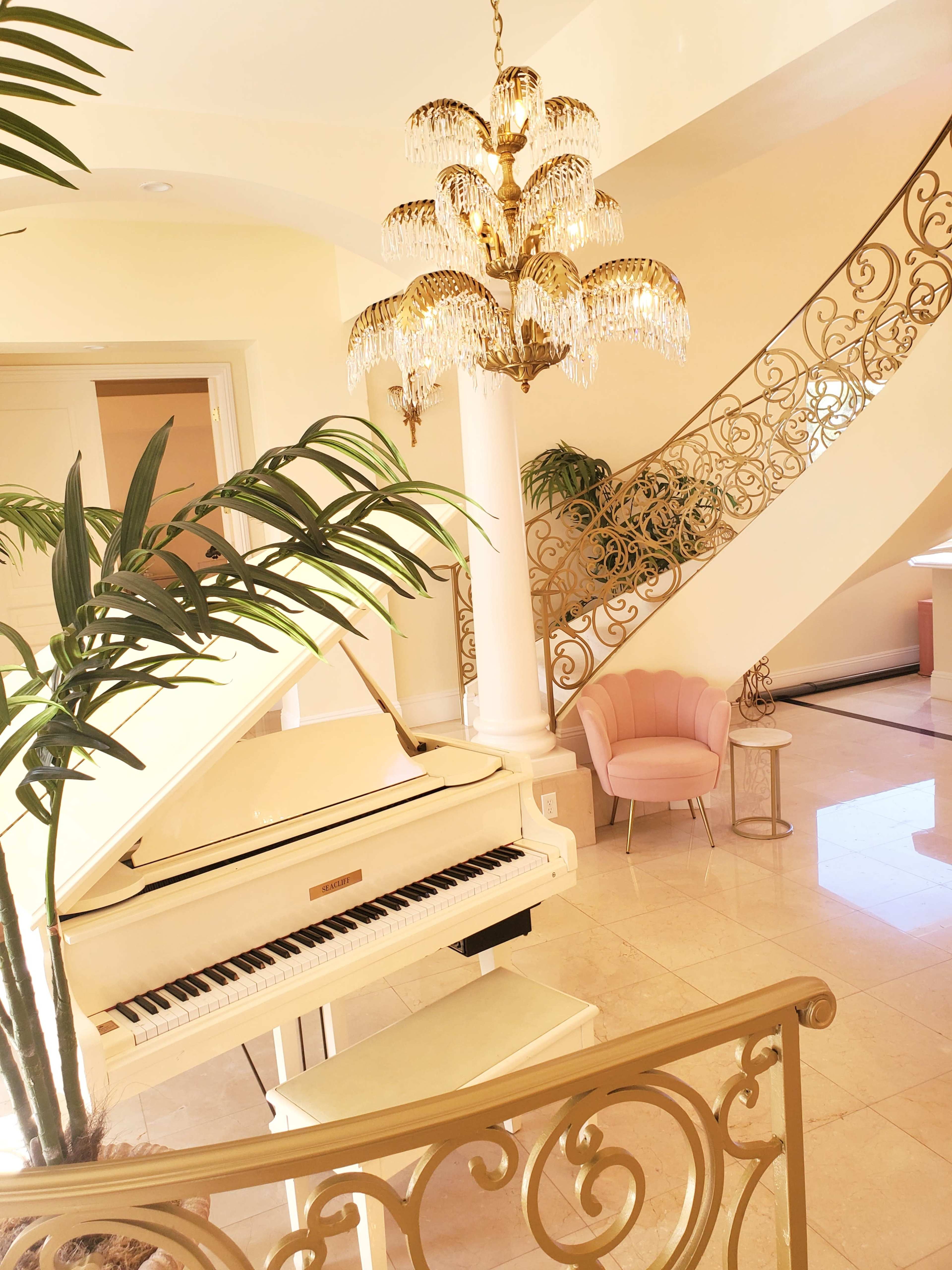 A grand piano sits beside a staircase adorned with ornate wrought iron, surrounded by indoor plants and a stylish chair in a brightly lit entryway.