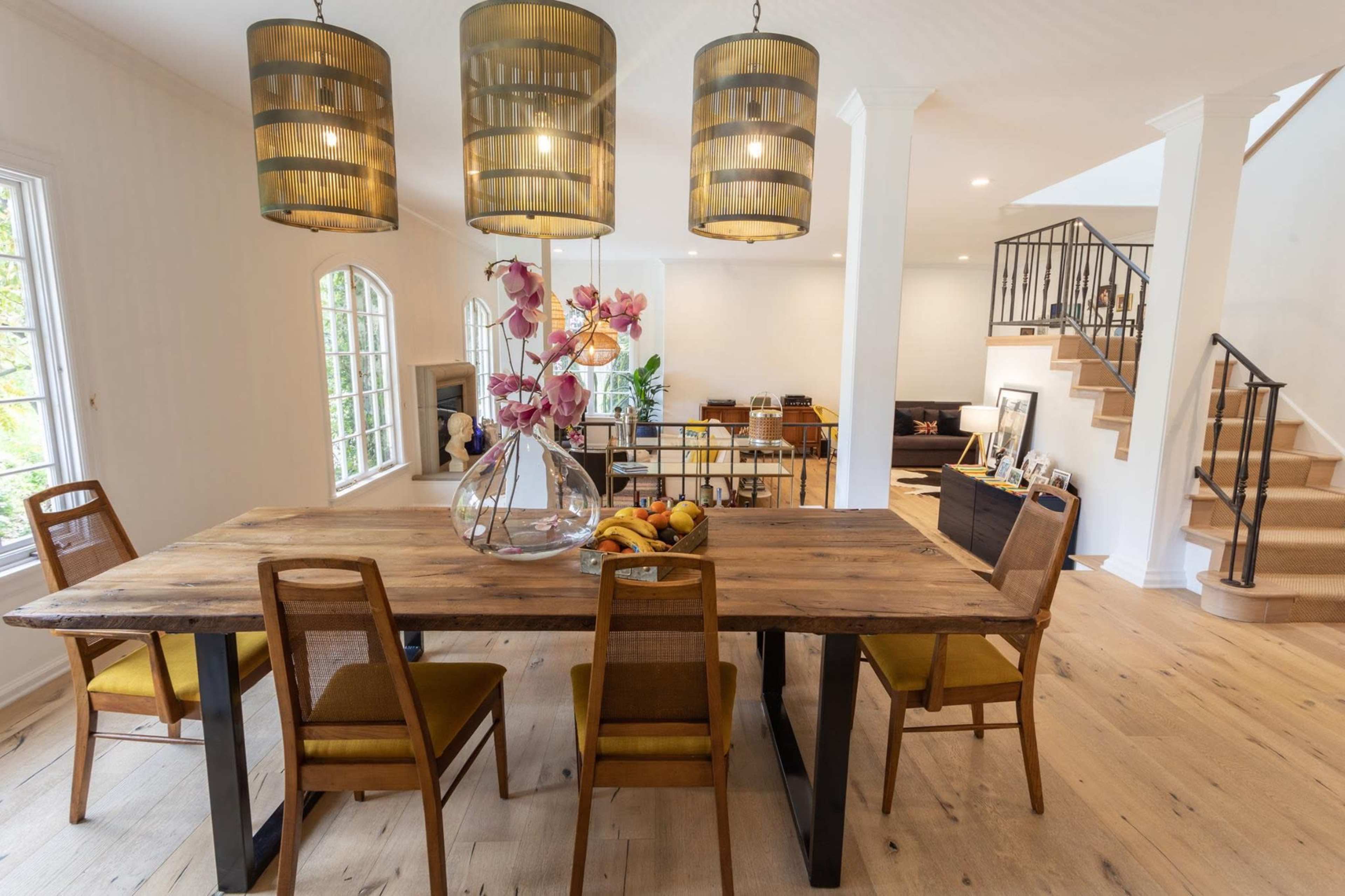 A wooden dining table with four chairs is set in a bright room adorned with three pendant lights and a floral centerpiece.