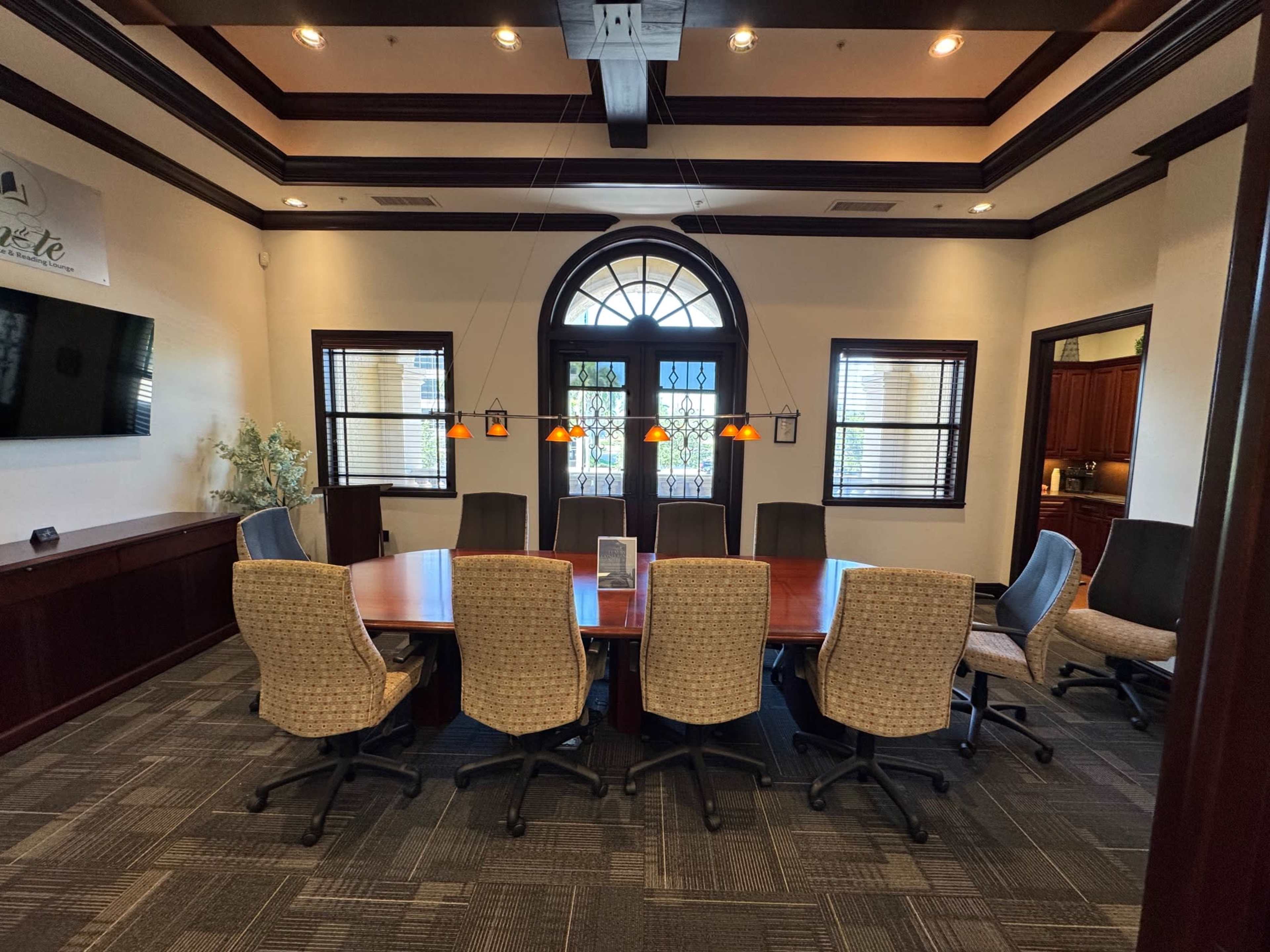 A large conference room features a rectangular wooden table surrounded by ten high-backed chairs, with pendant lights hanging above, and a wall-mounted screen on the left.