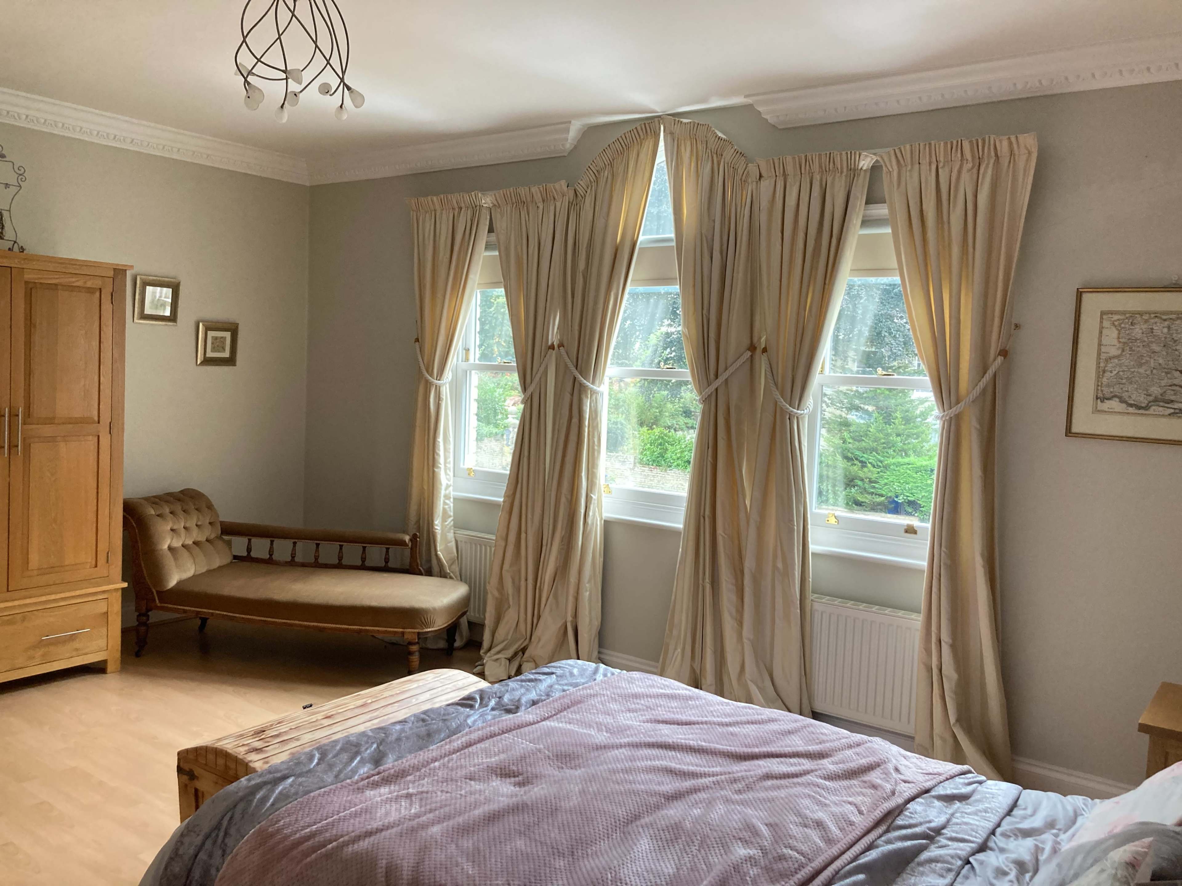 A bedroom features a bed with a pink coverlet, a wooden bench by the window, and long curtains framing large windows that overlook a garden.