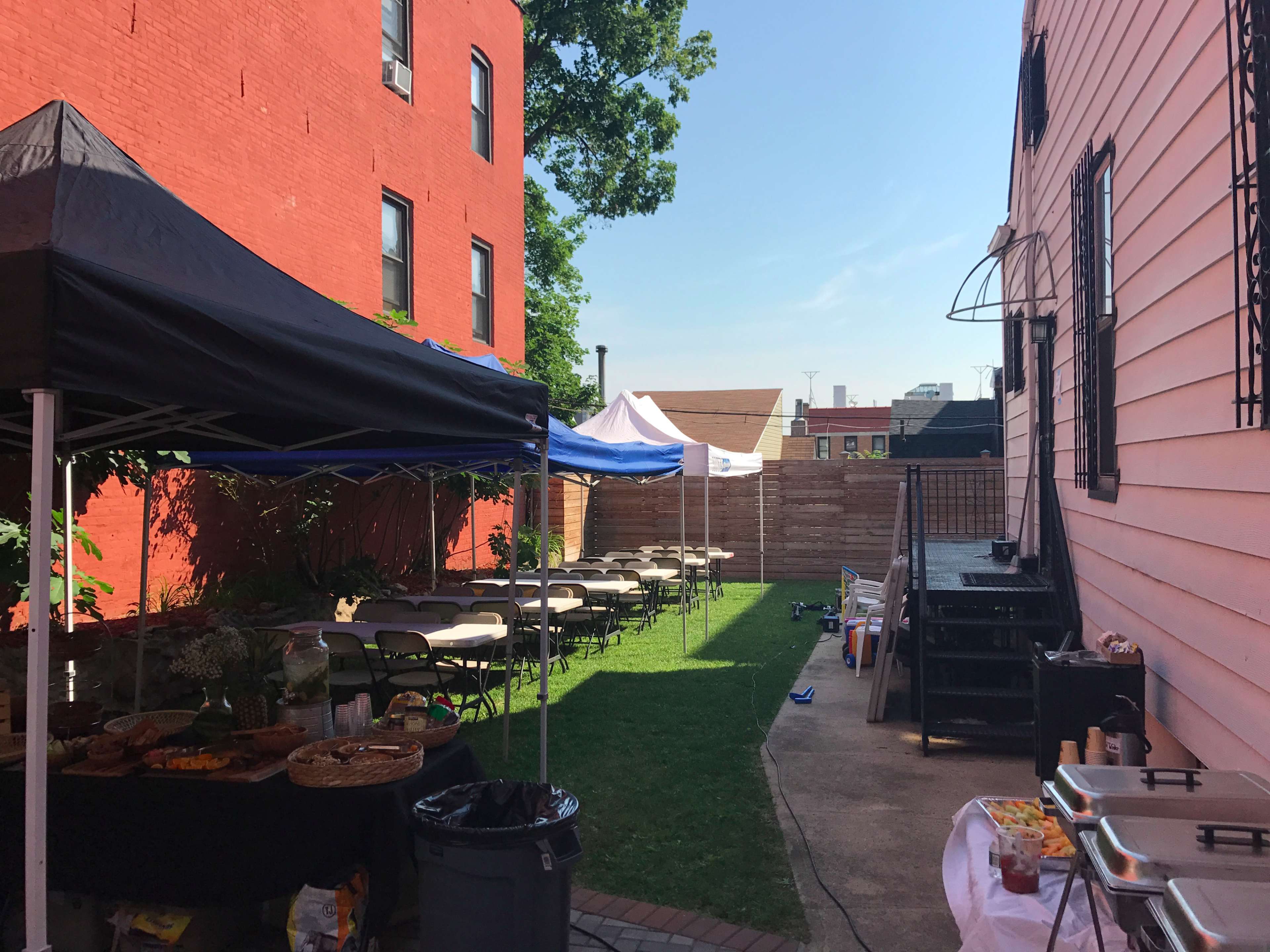A backyard space features tents, chairs, and tables set up for an outdoor gathering, with a grassy area and a wooden deck visible in the background.