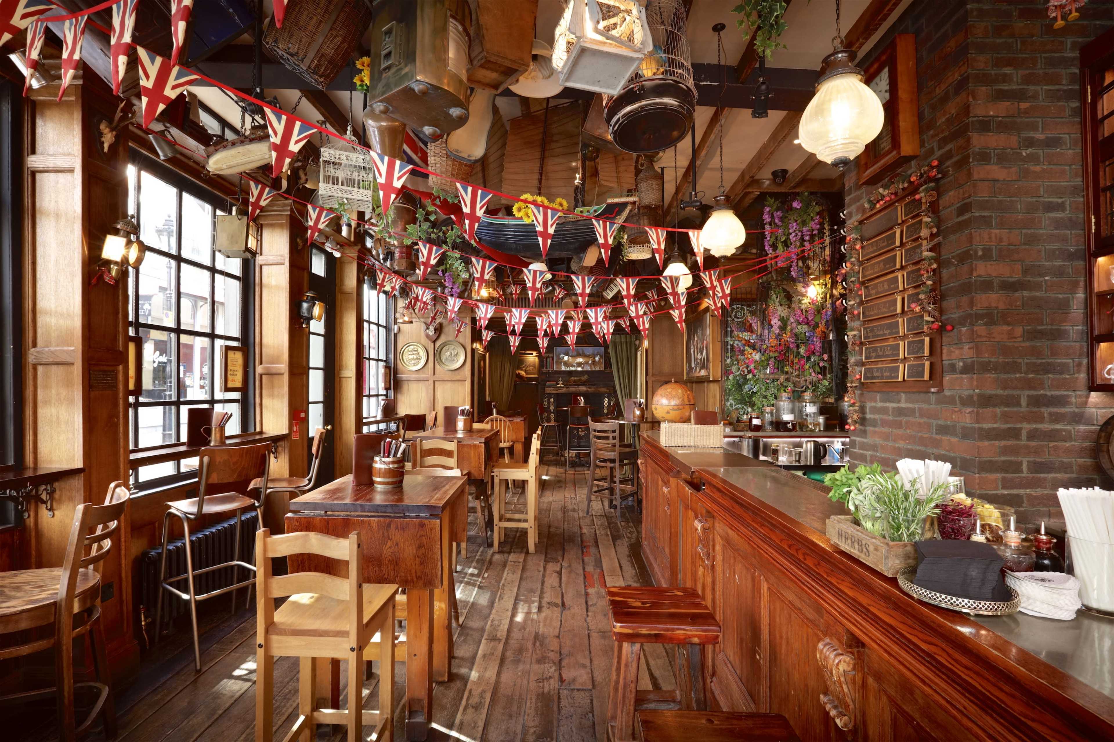 The image shows a rustic bar interior adorned with Union Jack bunting, wooden furniture, and a floral background.