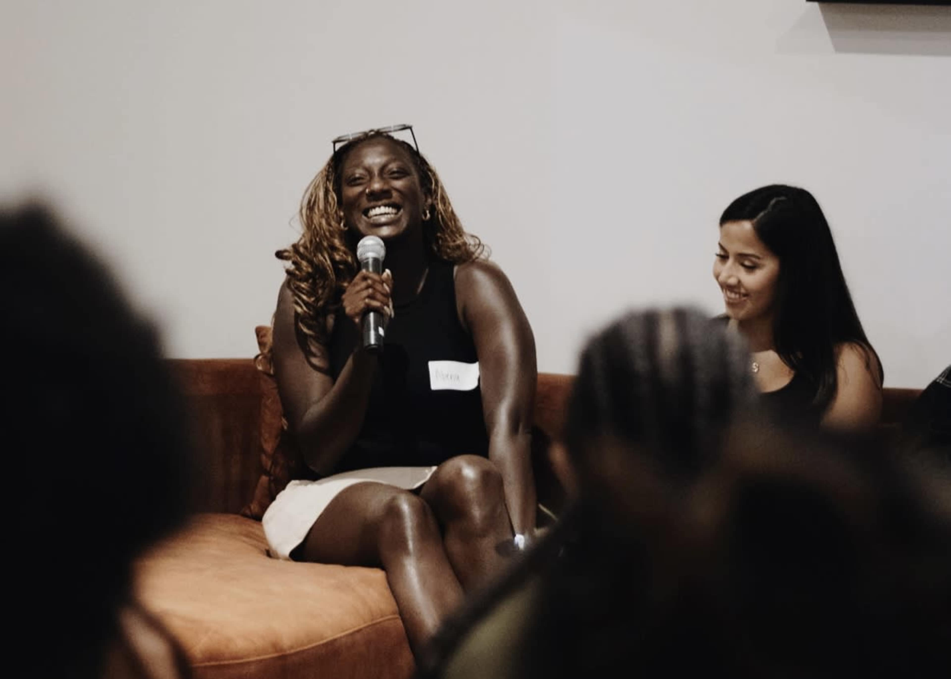 A woman wearing a black top and white skirt smiles while sitting on a couch and holding a microphone during a panel discussion.