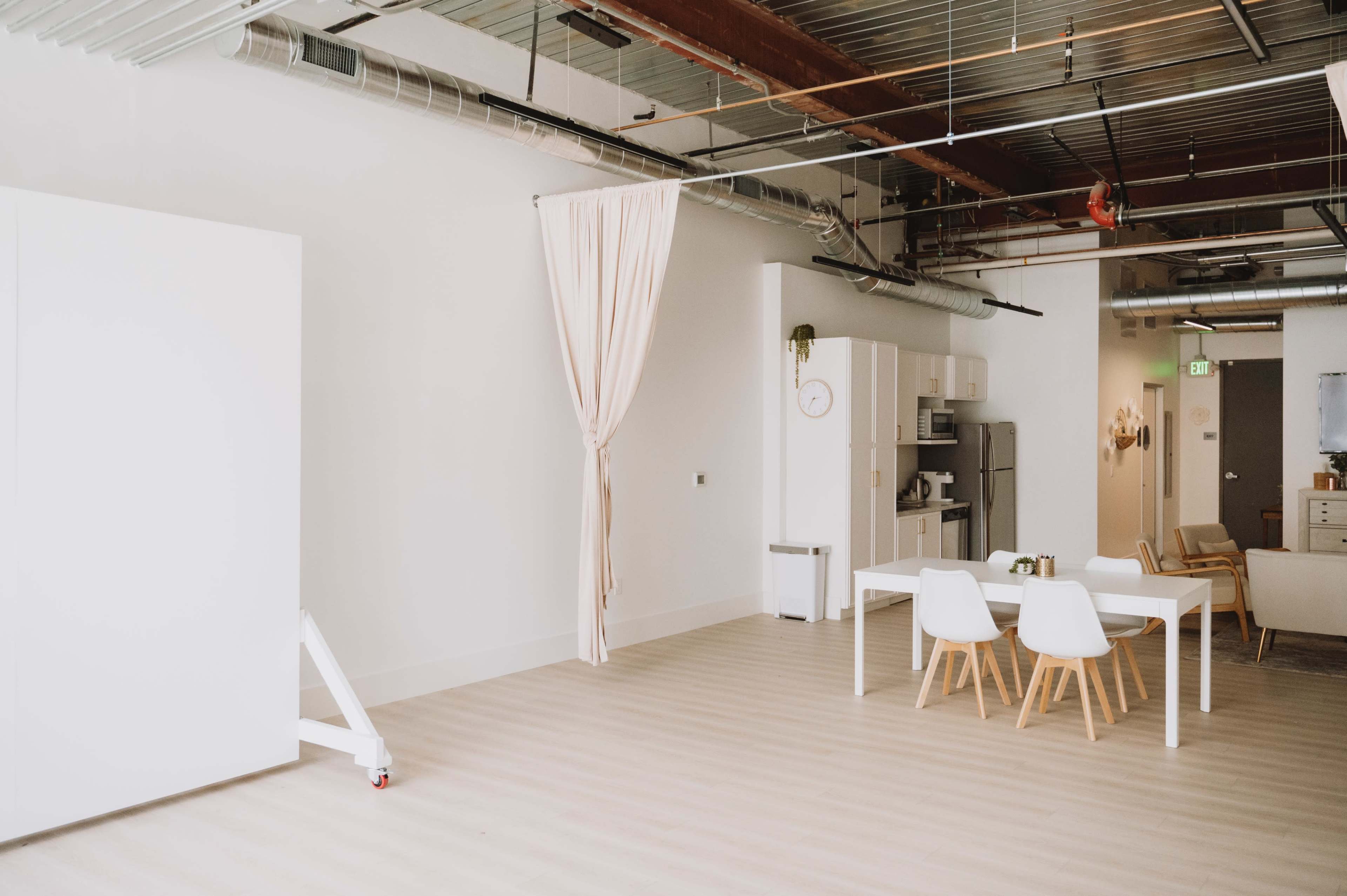 A minimalistic, bright interior space featuring a white kitchen setup, a dining table with chairs, and large windows, with a curtain partially drawn.