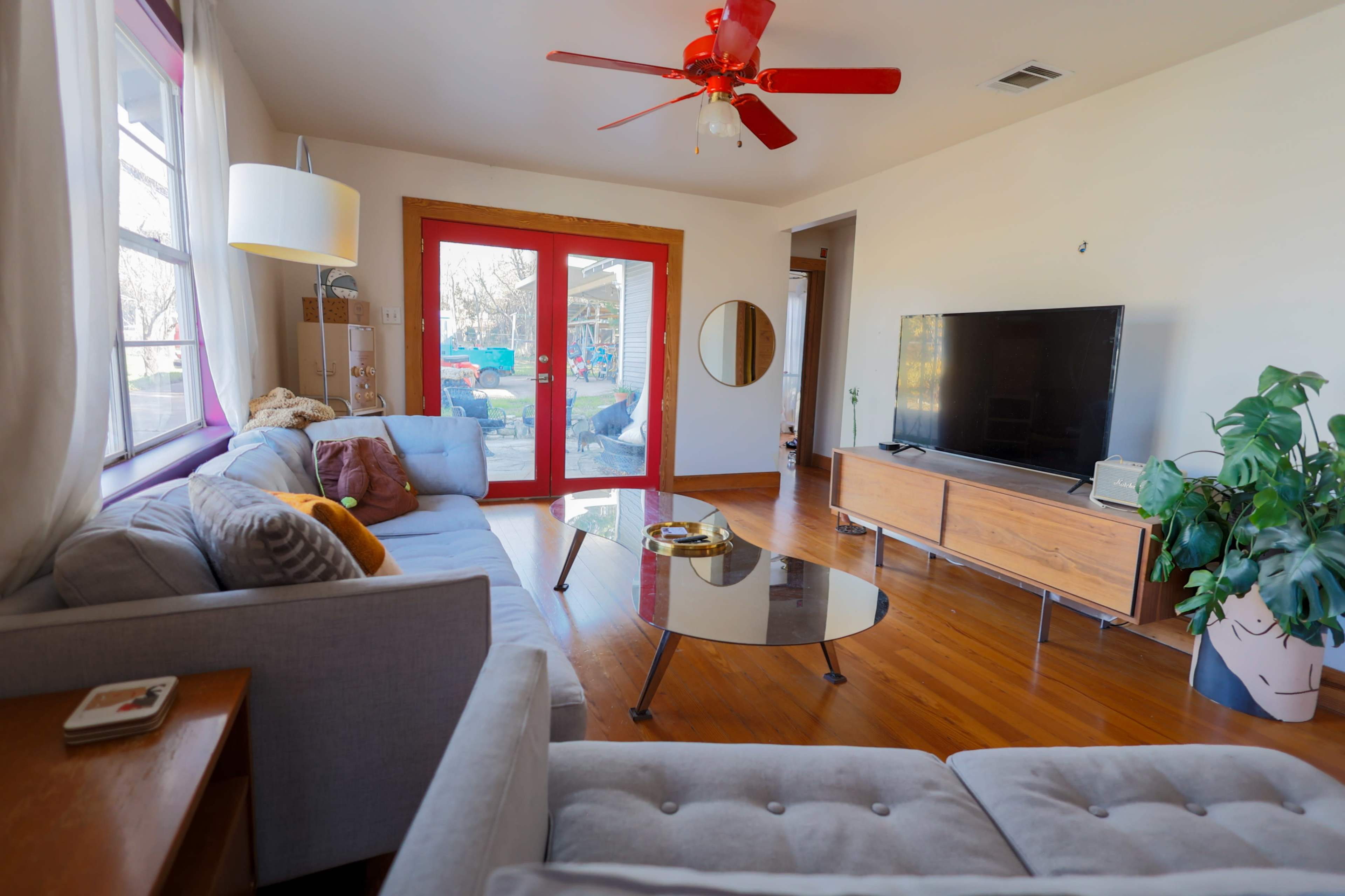 A bright living room features a gray sectional sofa, a glass coffee table, and a wooden media console beneath a large flat-screen TV, with red accents from the ceiling fan and window frames.