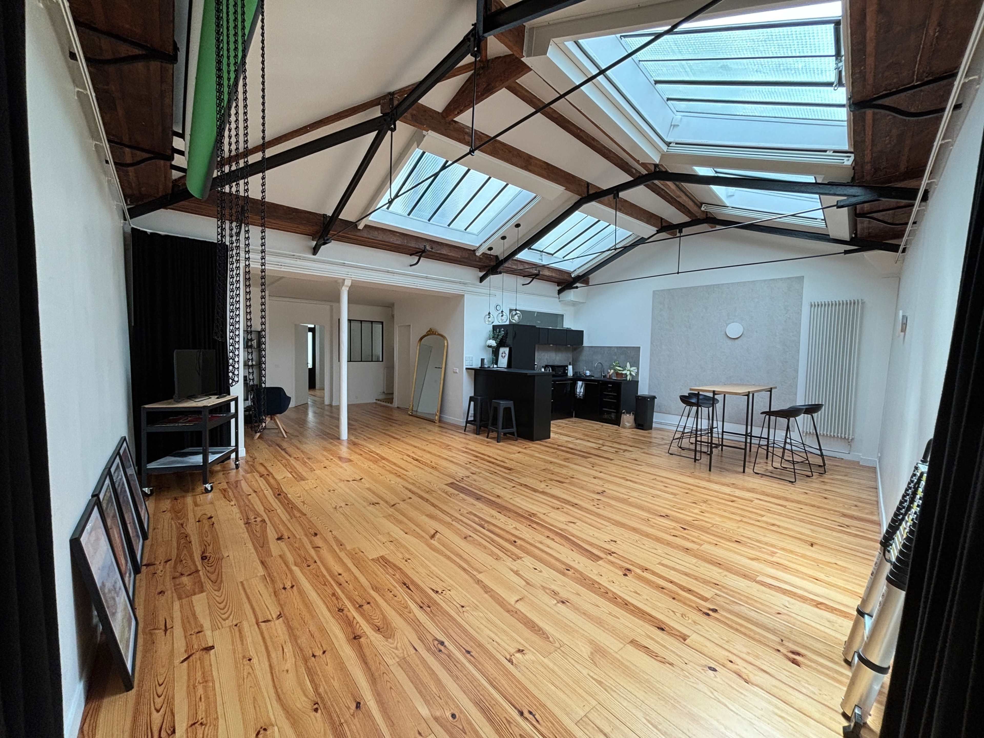 A spacious room with wooden flooring, large skylights, and various furniture arrangements including a kitchen area, tables, and chairs.