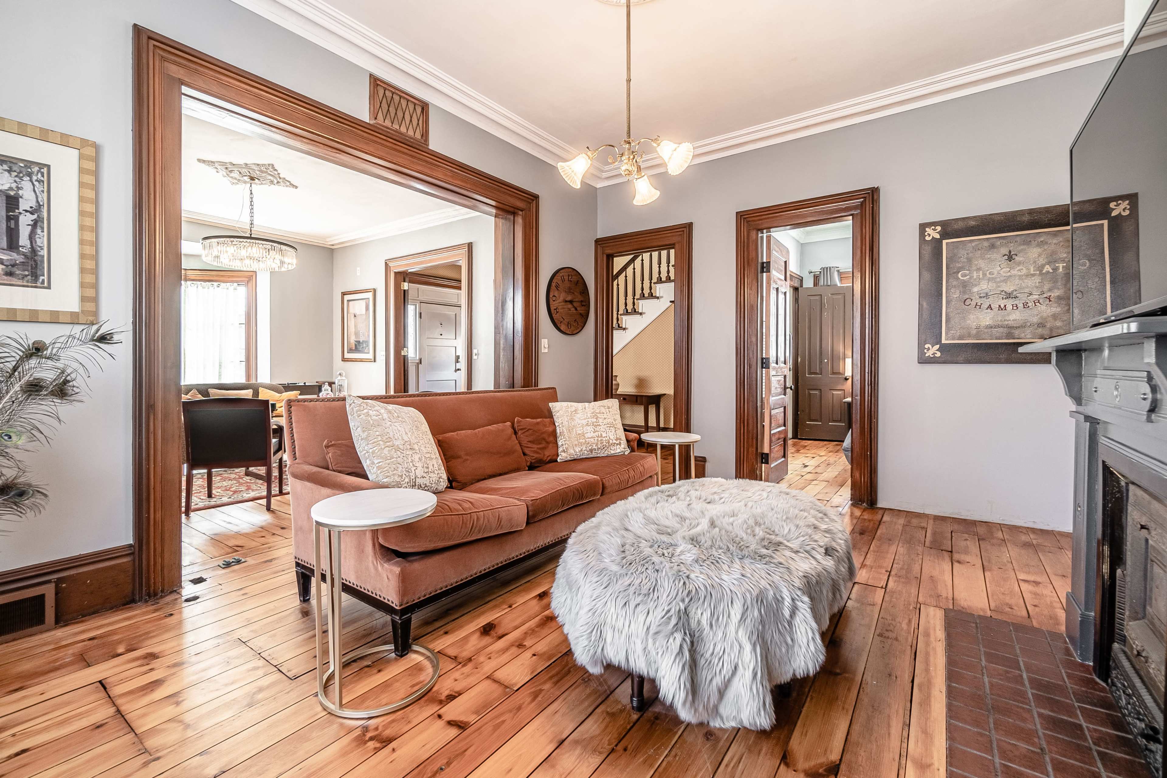 A living room with a brown sofa, a round fur ottoman, and an open view into a dining area with wooden flooring and classic architectural details.