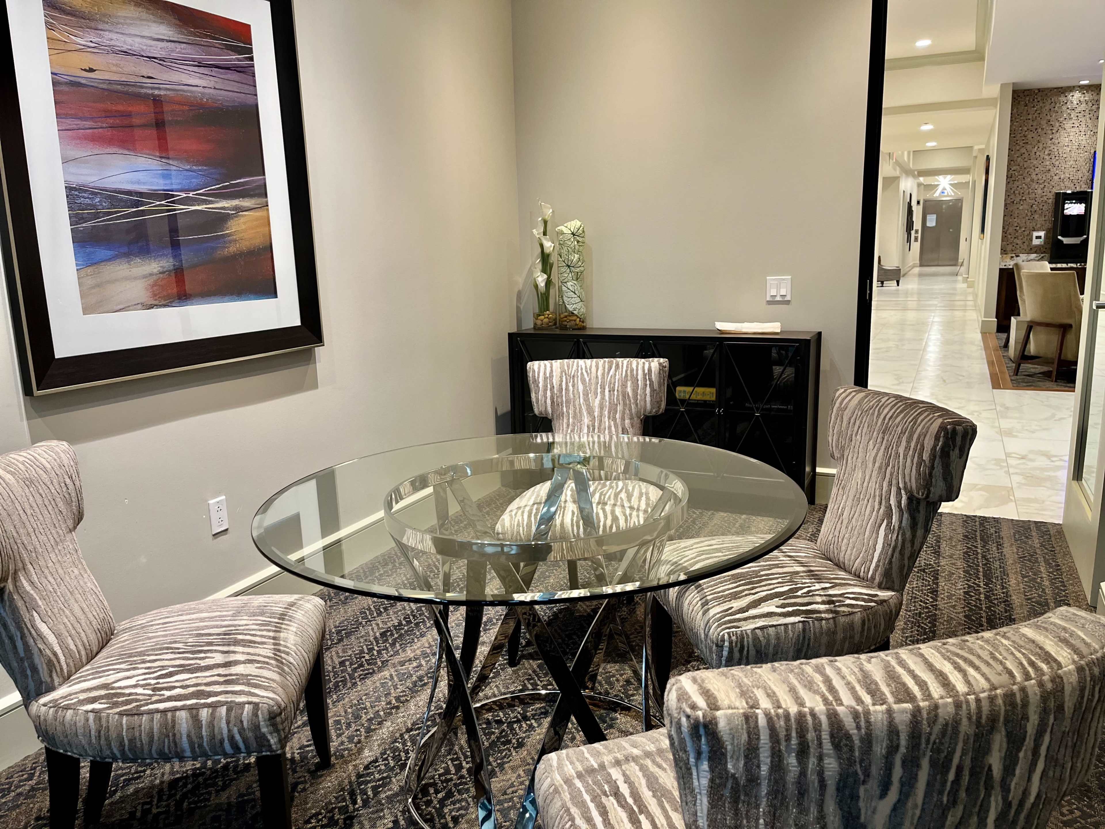 A round glass dining table is surrounded by four striped chairs in a well-lit room with art on the wall.