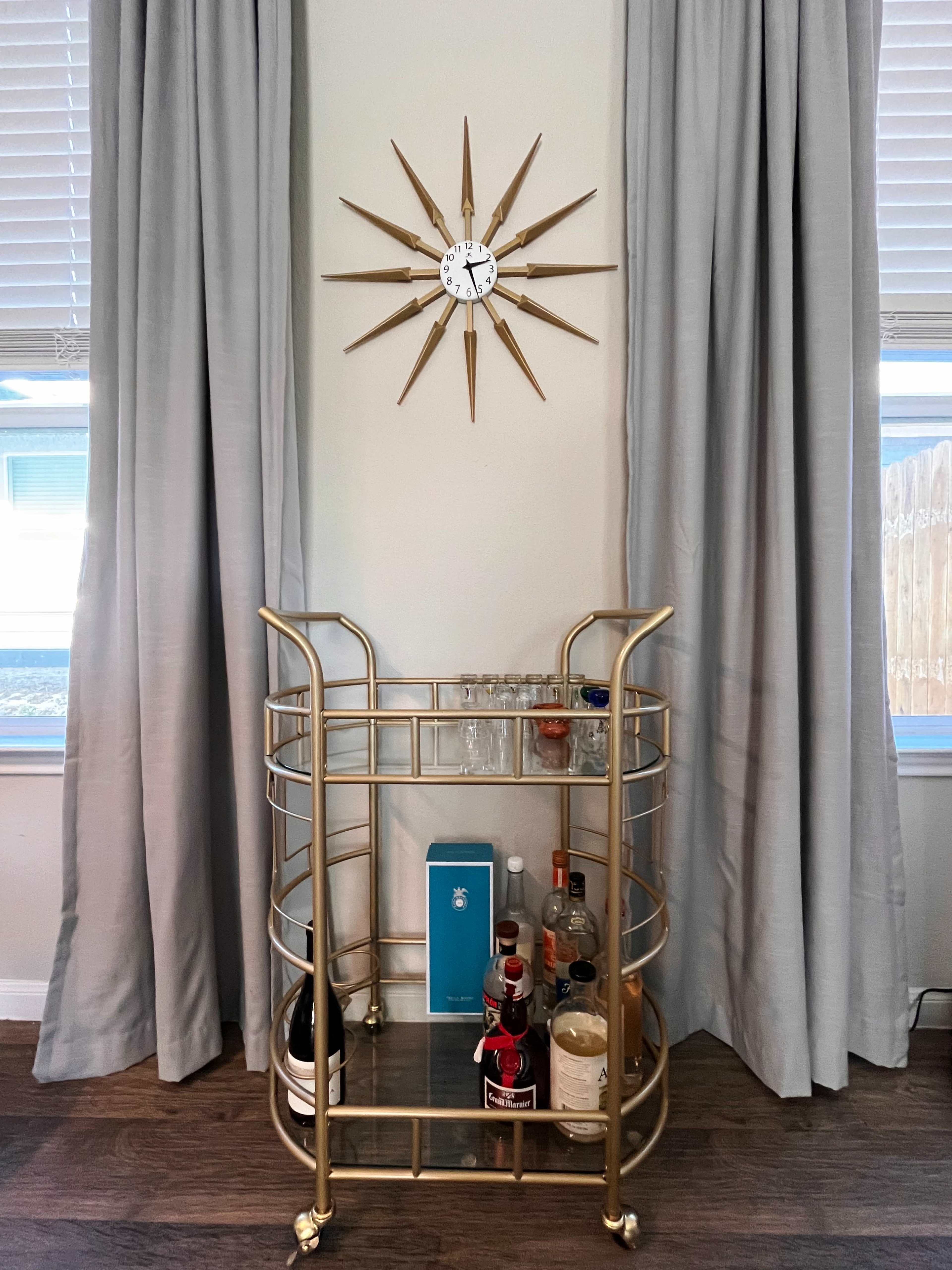 A gold bar cart with various bottles and glasses sits against a light-colored wall, topped by a starburst clock.