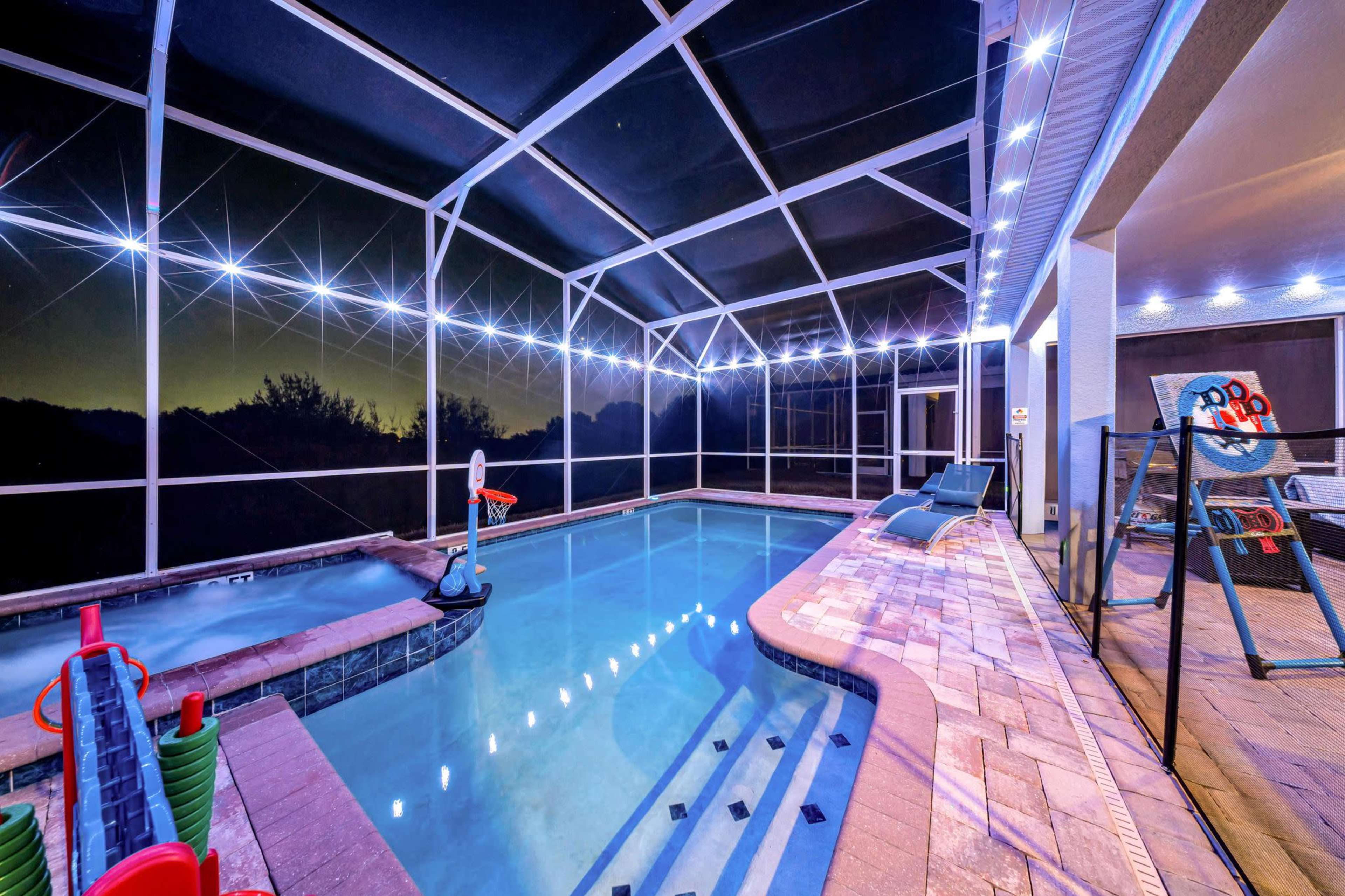 The image shows an illuminated indoor swimming pool surrounded by a screened enclosure, with a basketball hoop and lounge chairs nearby.