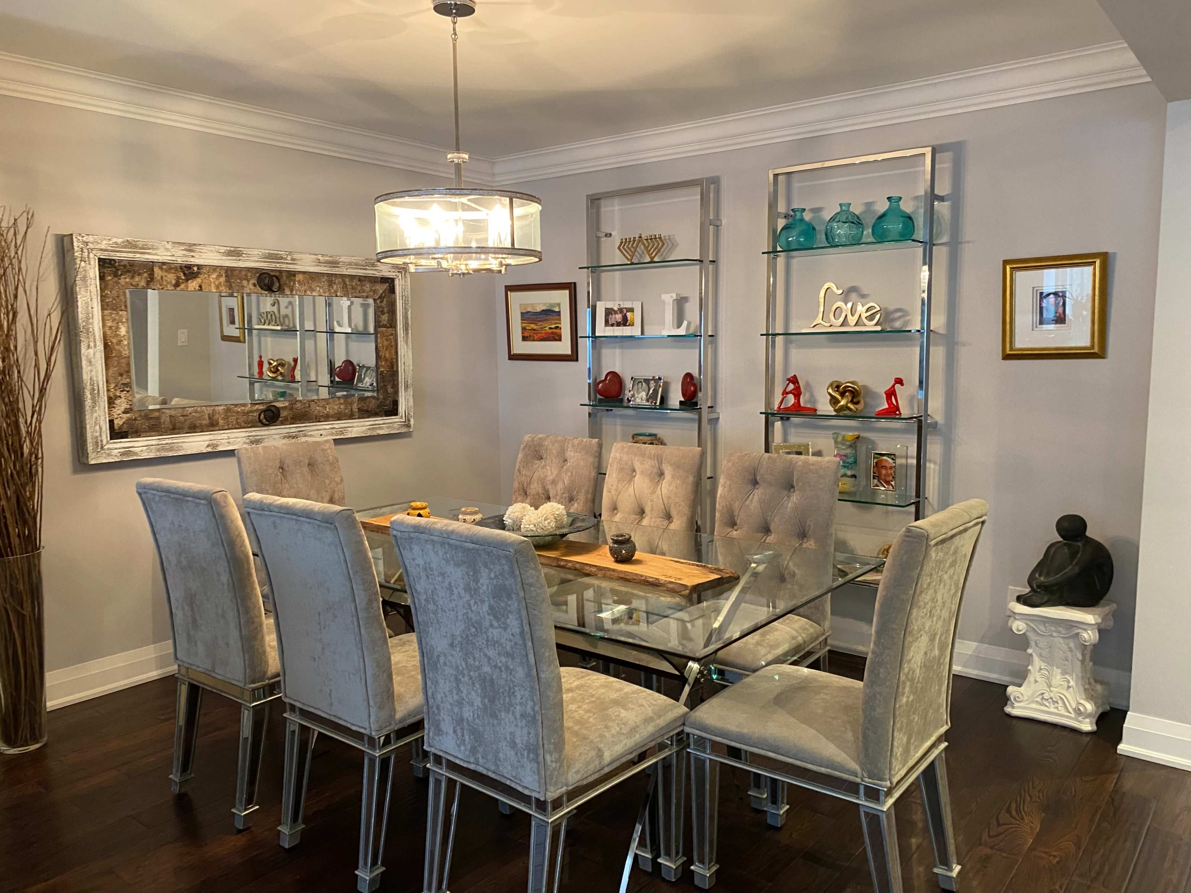 A modern dining room features a glass table surrounded by upholstered chairs, with a decorative mirror and shelves displaying various objects on the walls.