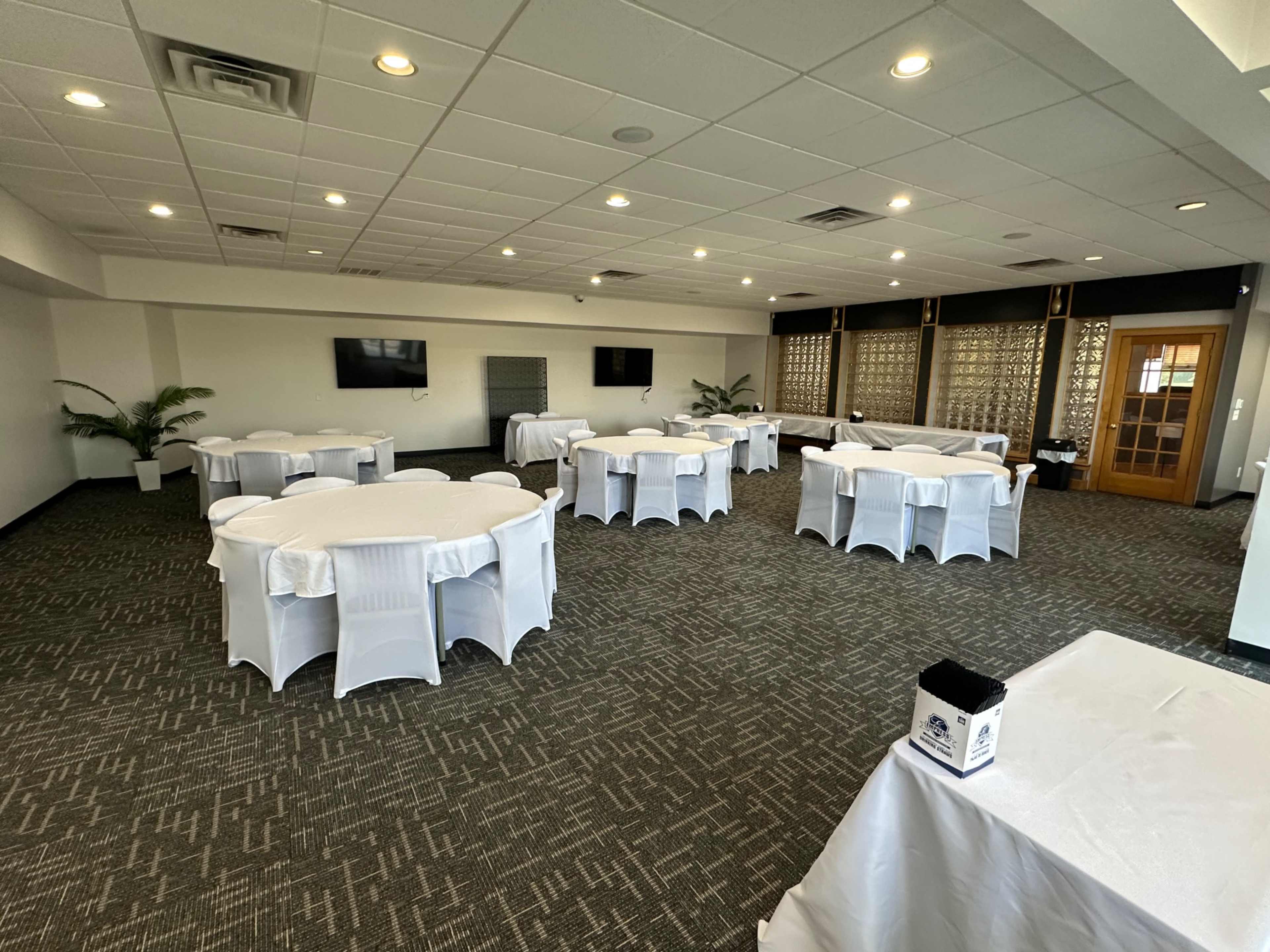 The image shows a spacious event room with round tables covered in white linens and equipped with chairs, featuring two television screens and decorative paneling.