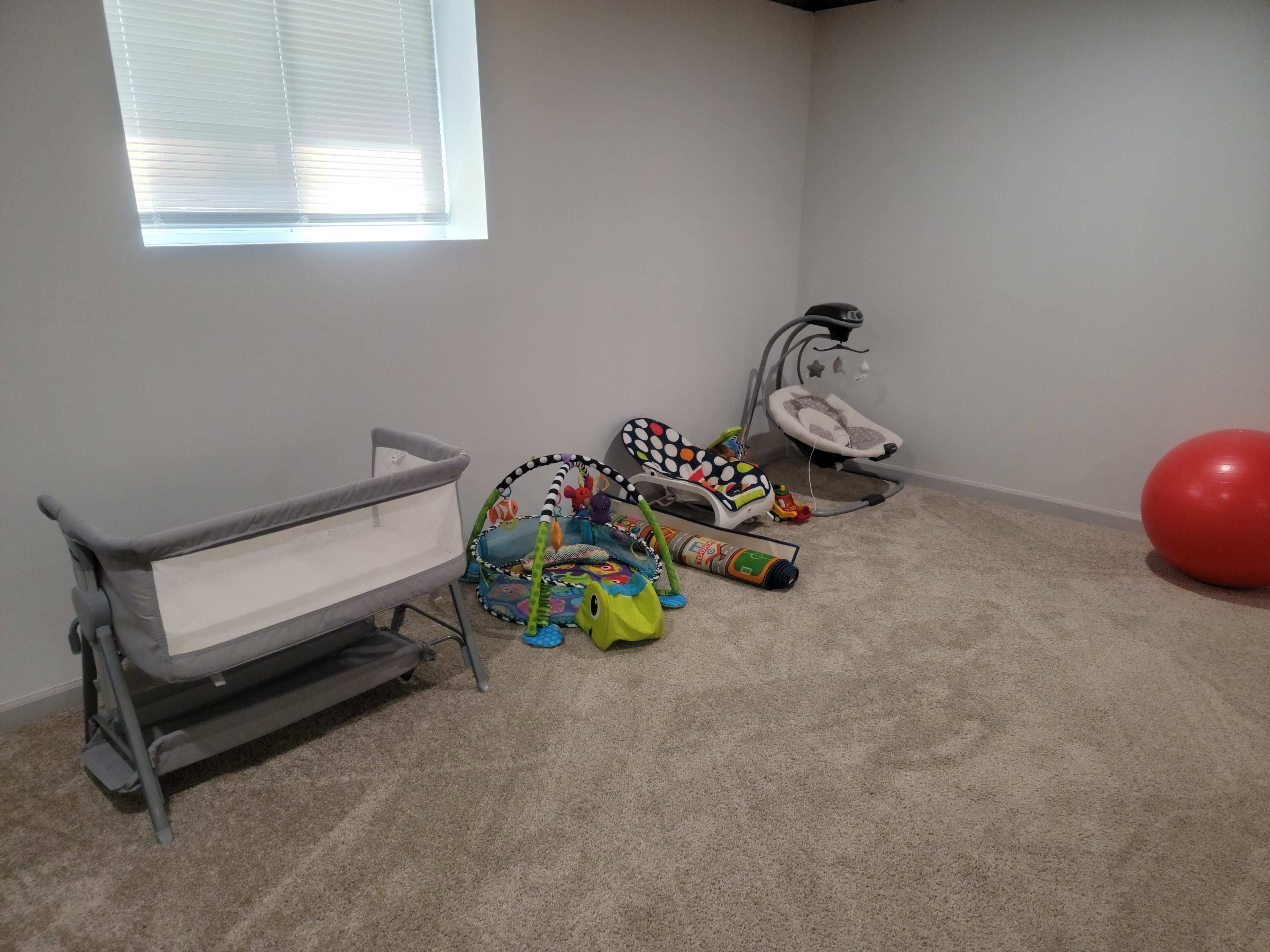 A room with a light-colored carpet, featuring a bassinet, various colorful baby toys, a baby seat, and a large red exercise ball.