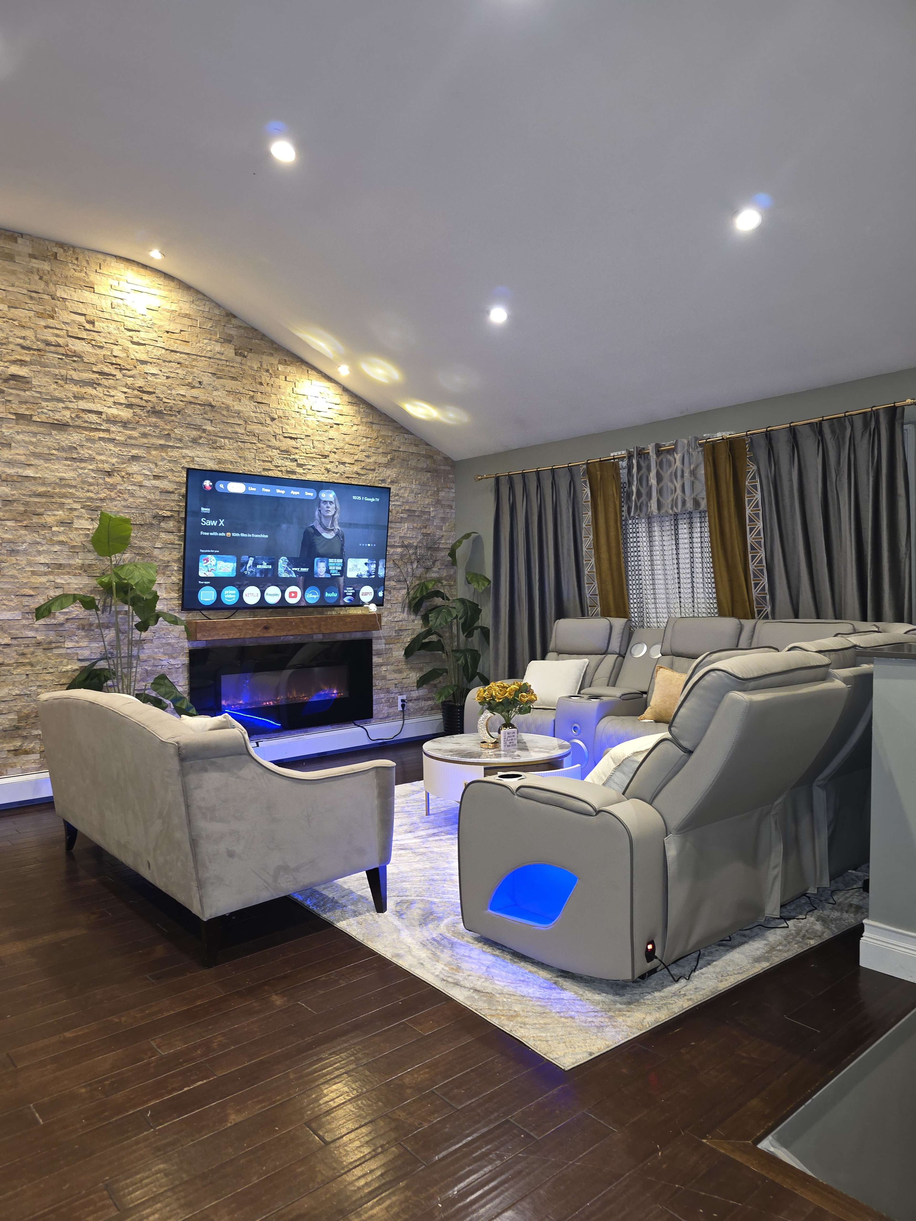 The image shows a modern living room with a stone accent wall, a large TV, comfortable seating, and decorative plants.