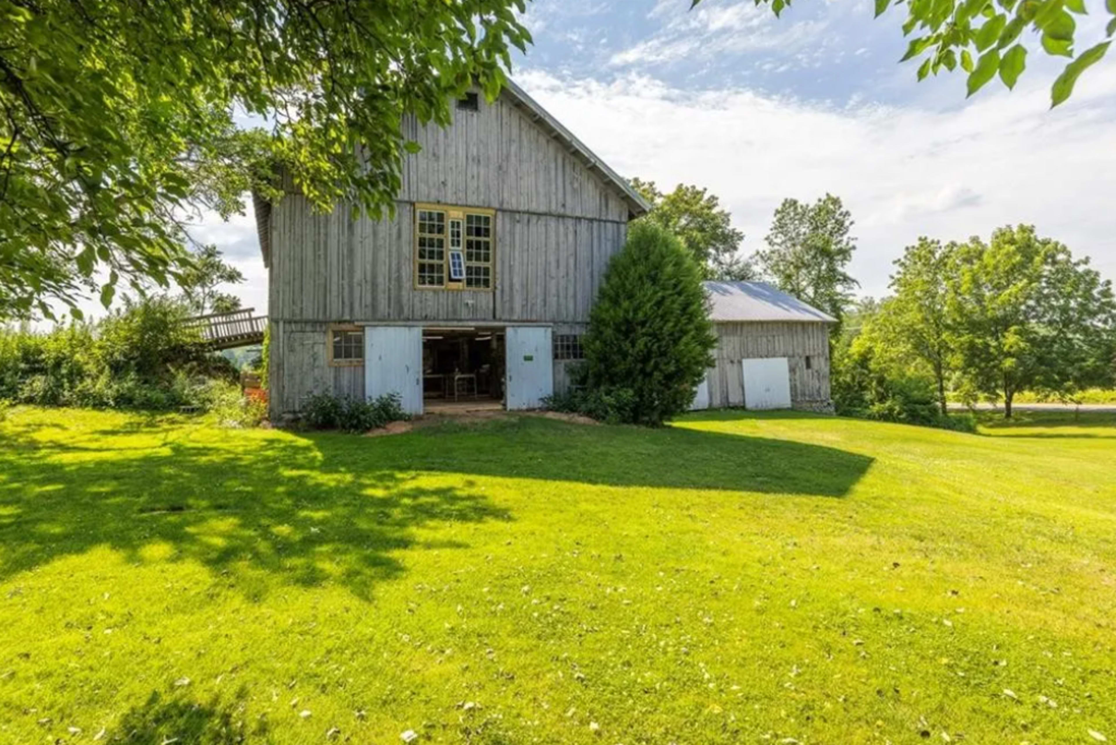 A wooden barn stands amidst green grass and trees under a partly cloudy sky.