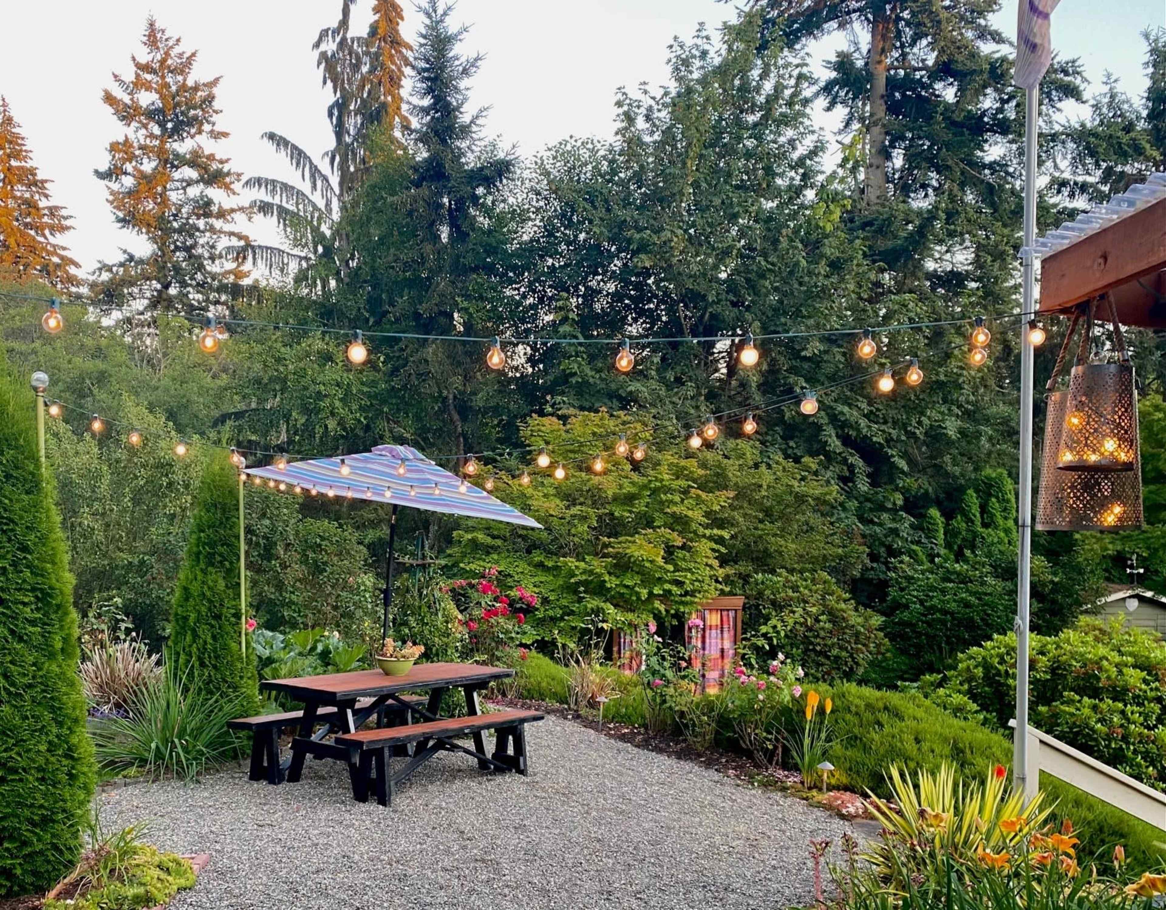 A gravel patio features a picnic table under a striped umbrella, surrounded by garden plants and strung lights overhead.